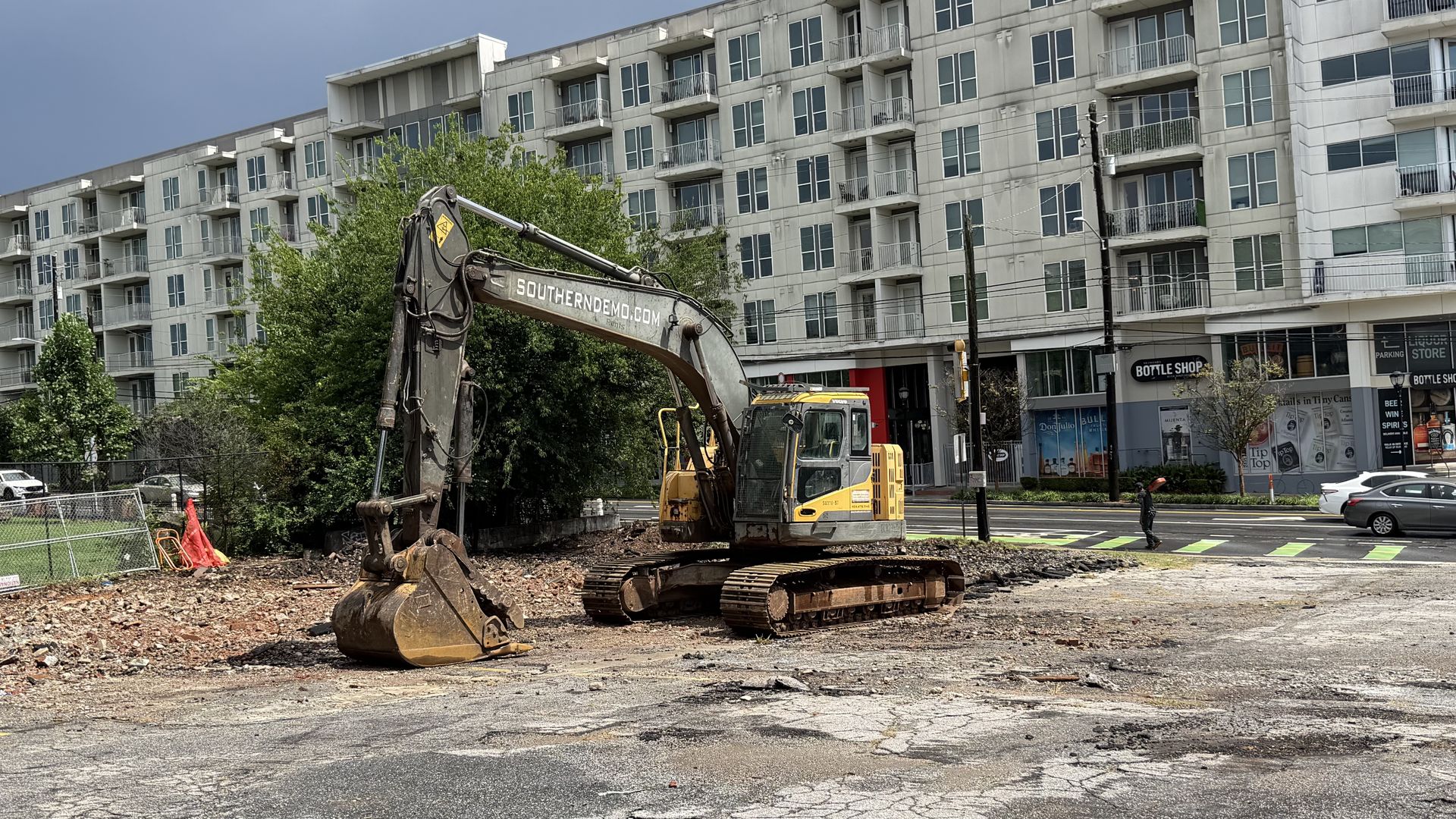 Yellow excavator on a rubble-strewn construction site in front of a gray, multi-story apartment building with balconies; a Bottle Shop storefront and cars line the street.
