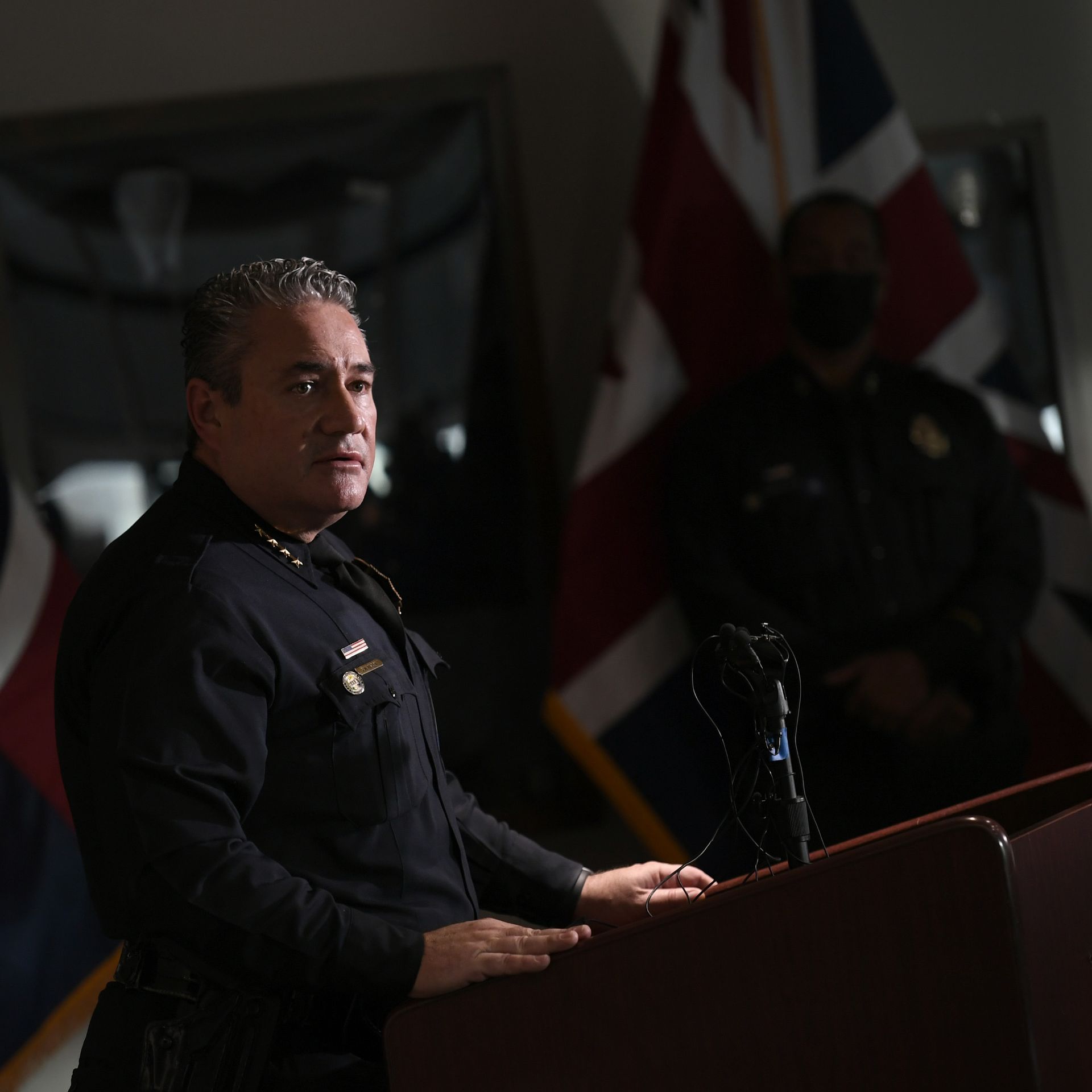 A police chief in full uniform stands near a lectern in a dimly-lit room while addressing member of the press.  