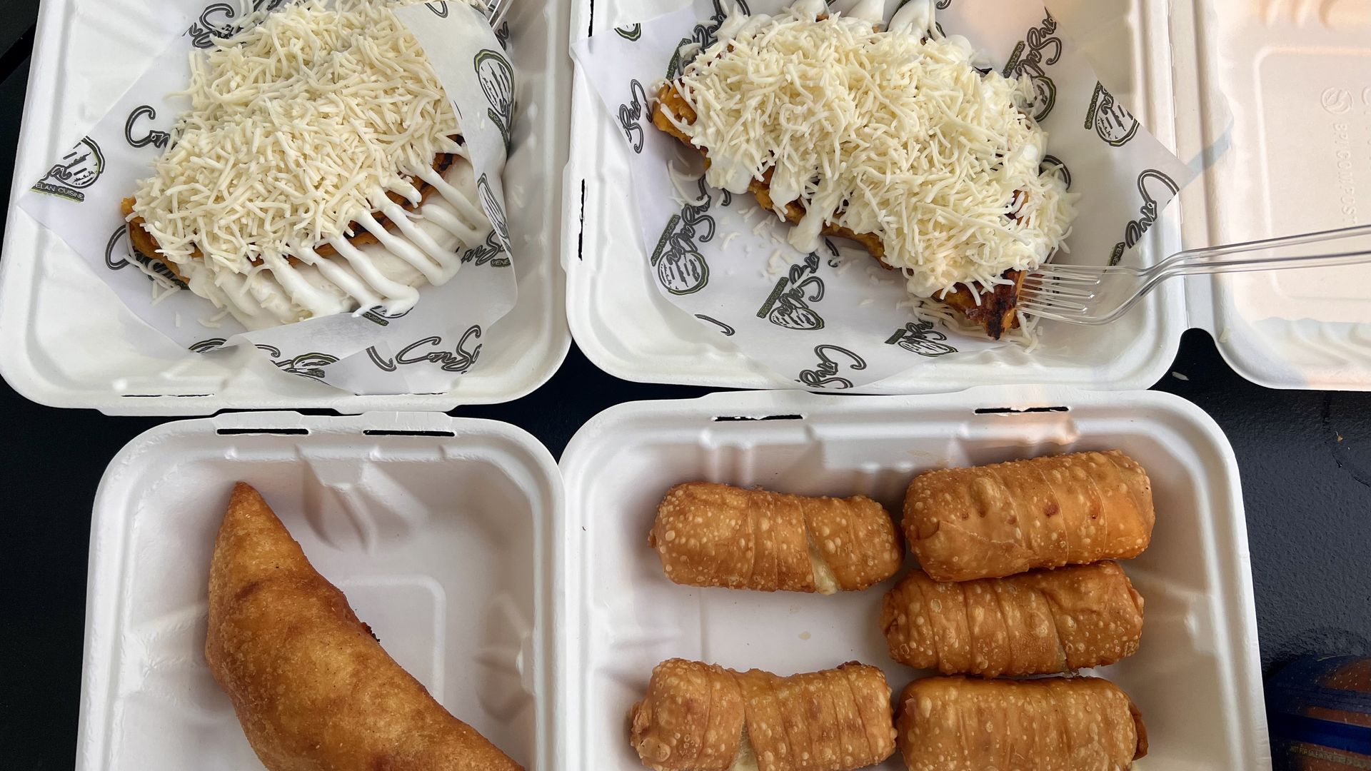 Four styrofoam containers with food: two with cheese-covered dishes topped with white sauce, one with a fried empanada, and one with five fried cheese sticks.
