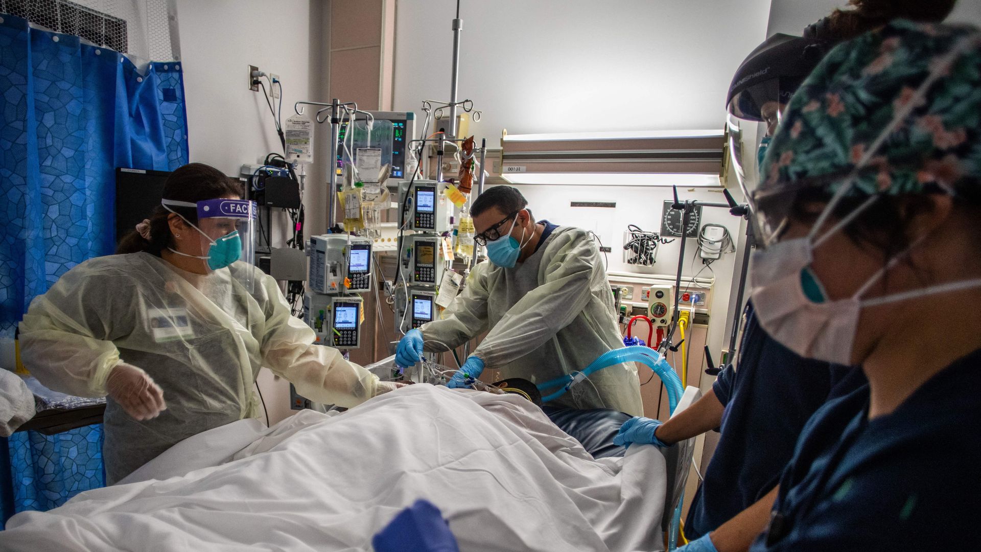 Health care workers in masks and protective gear care for a COVID-19 patient on a hospital bed.