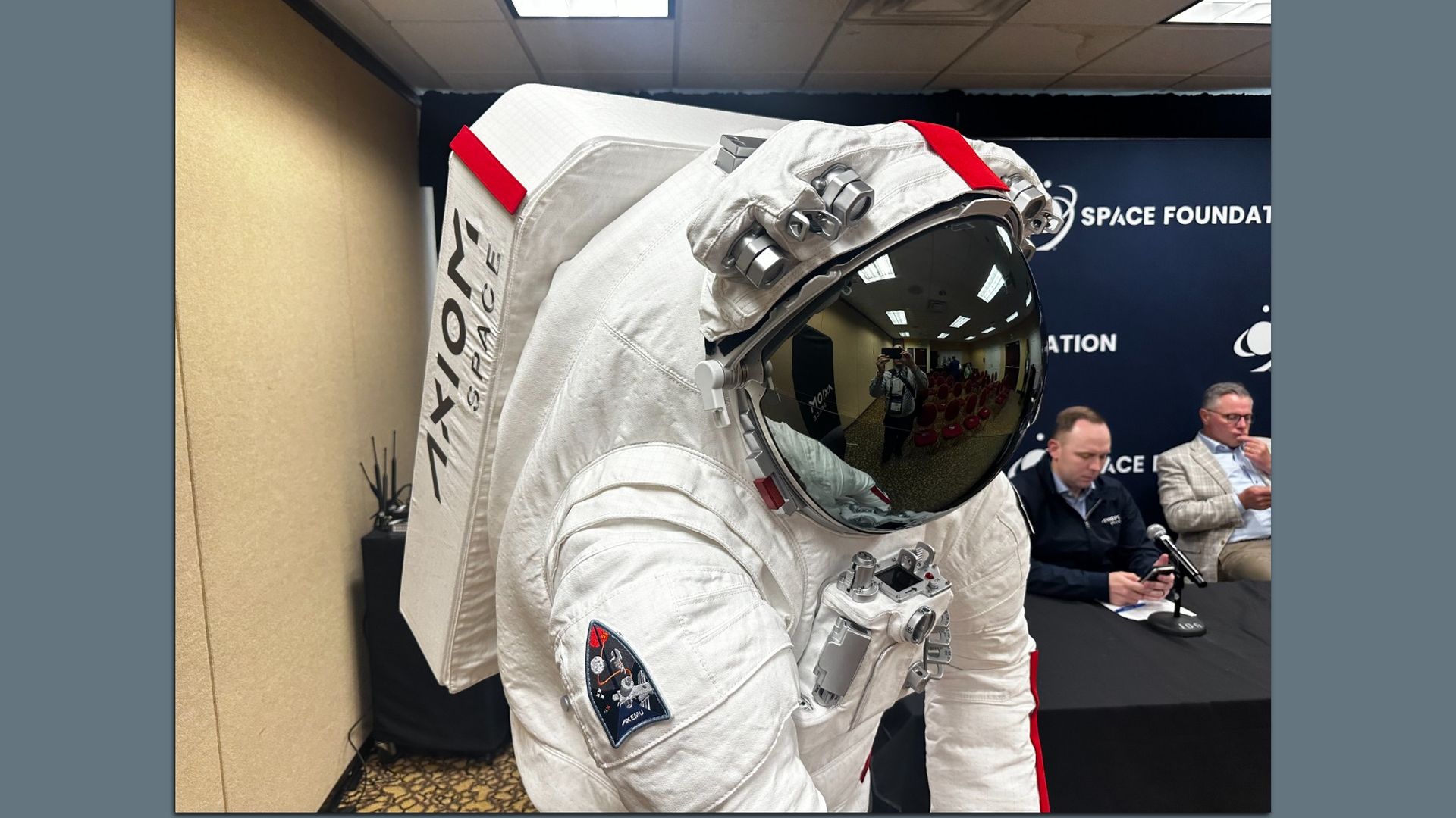Foreground shows a white spacesuit with a reflective visor and red trim; in the background a Space Foundation banner and three men seated at a panel table in a conference room.