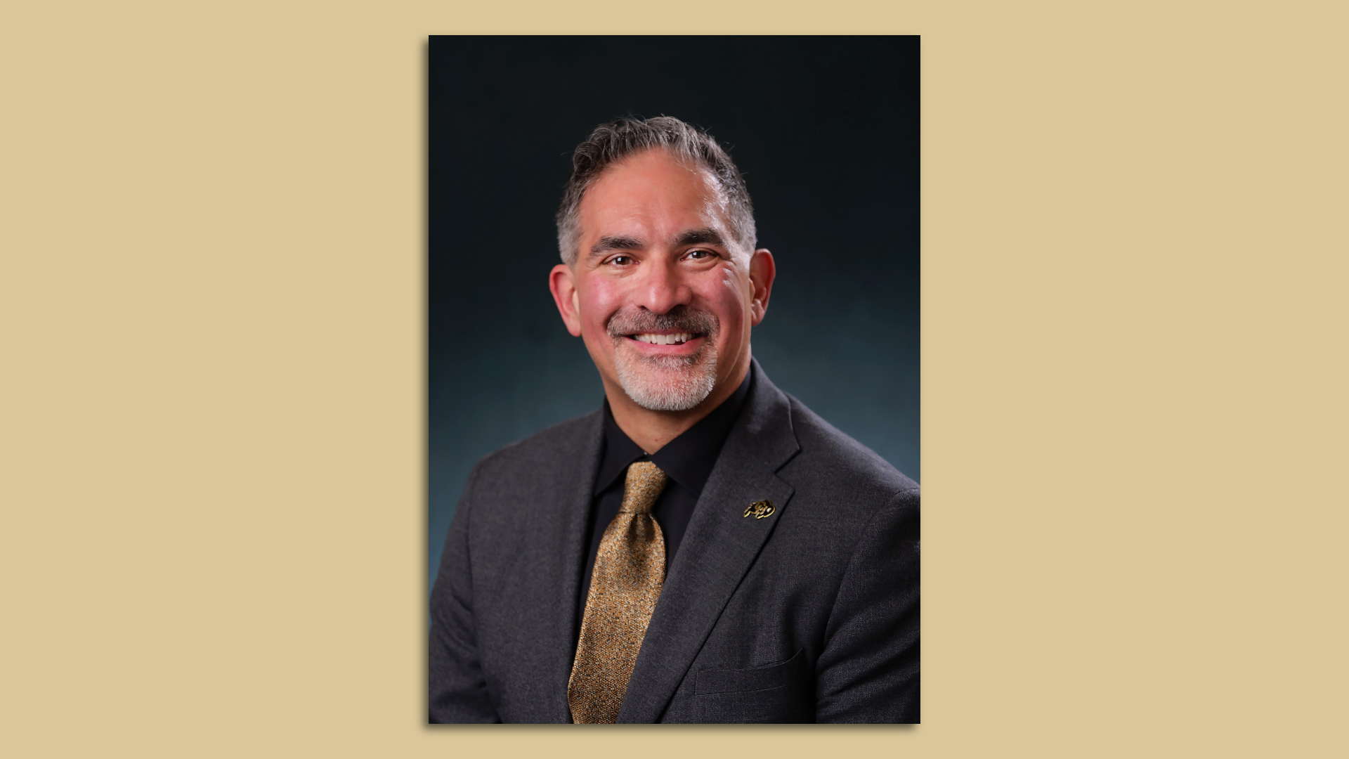 A man smiles for a portrait. He is wearing a dark gray jacket, a black shirt and a gold necktie. 
