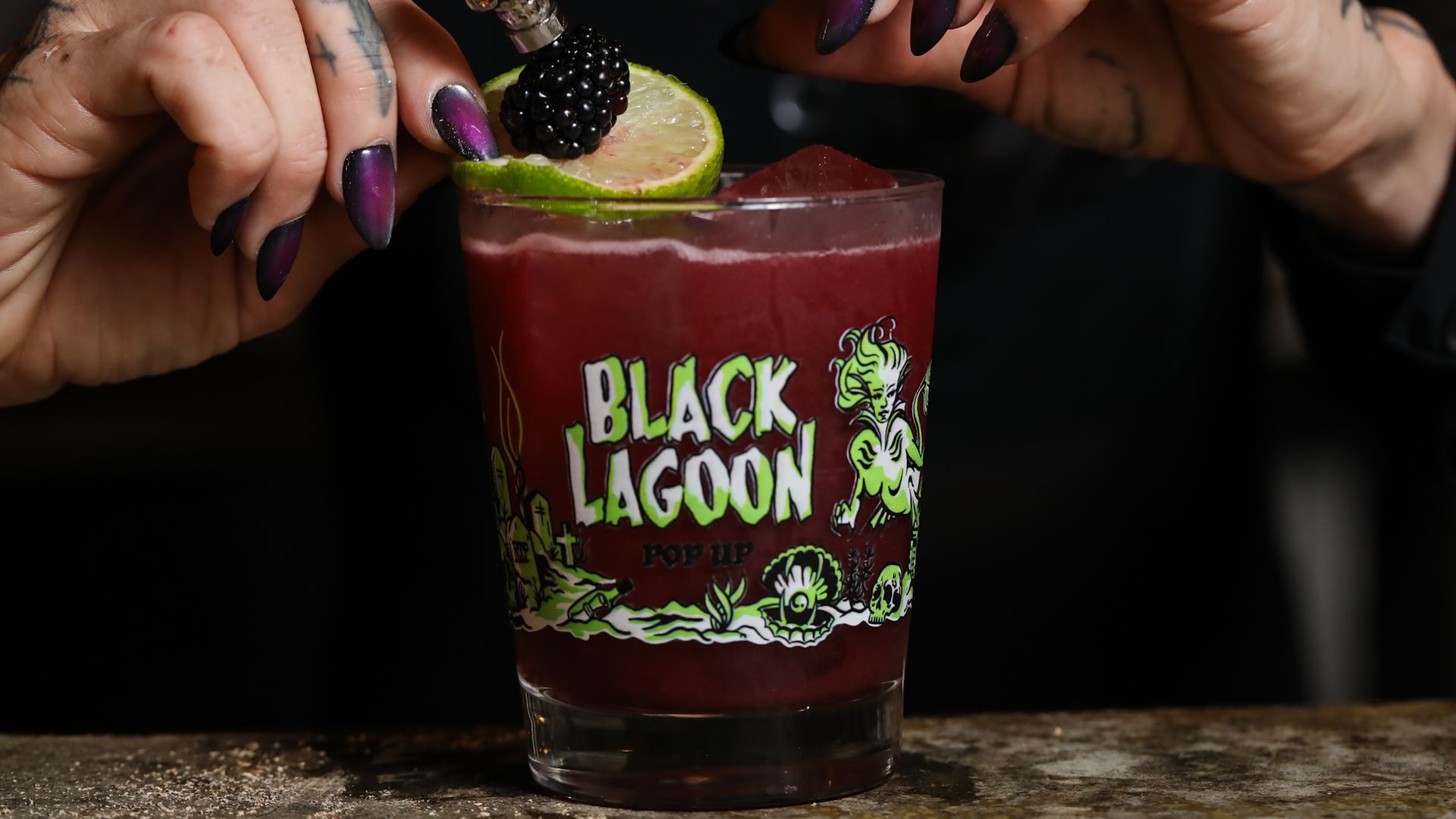 Close-up of a red cocktail in a cup labeled "Black Lagoon," garnished with a lime slice and blackberry, held by tattooed hands with purple-to-black gradient nails; a skull-topped bottle rests beside.