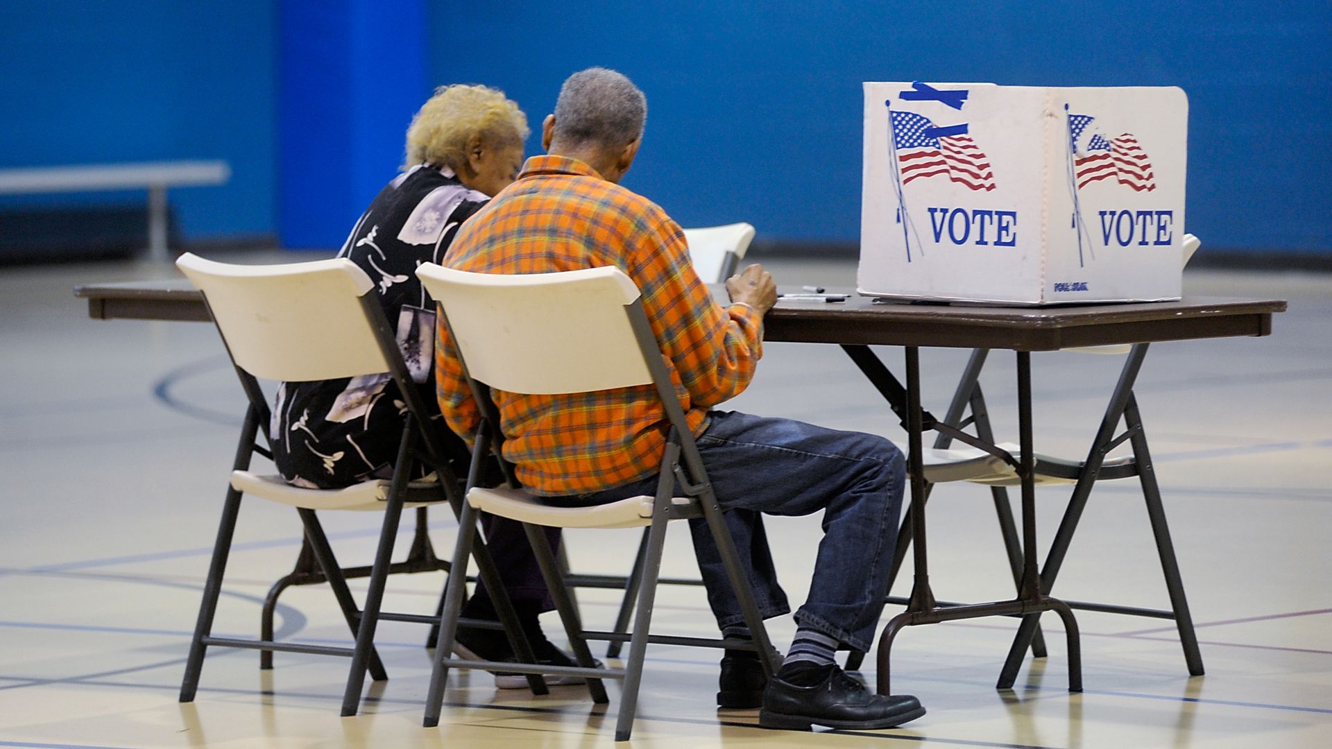 Two people sit side by side at a table in a polling place, filling out their ballots behind voting privacy screens decorated with American flags and the word “VOTE.”