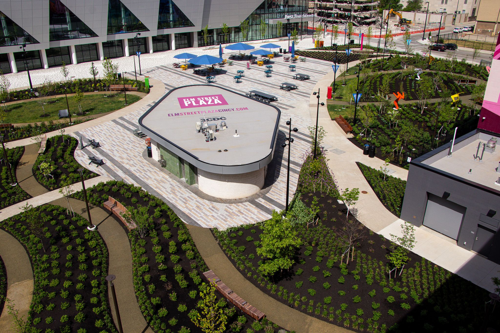 An aerial view of Elm Street Plaza: a modern glass-front building, a tiled plaza with blue umbrellas, a curved structure labeled "Elm Street Plaza," and manicured green gardens with benches.
