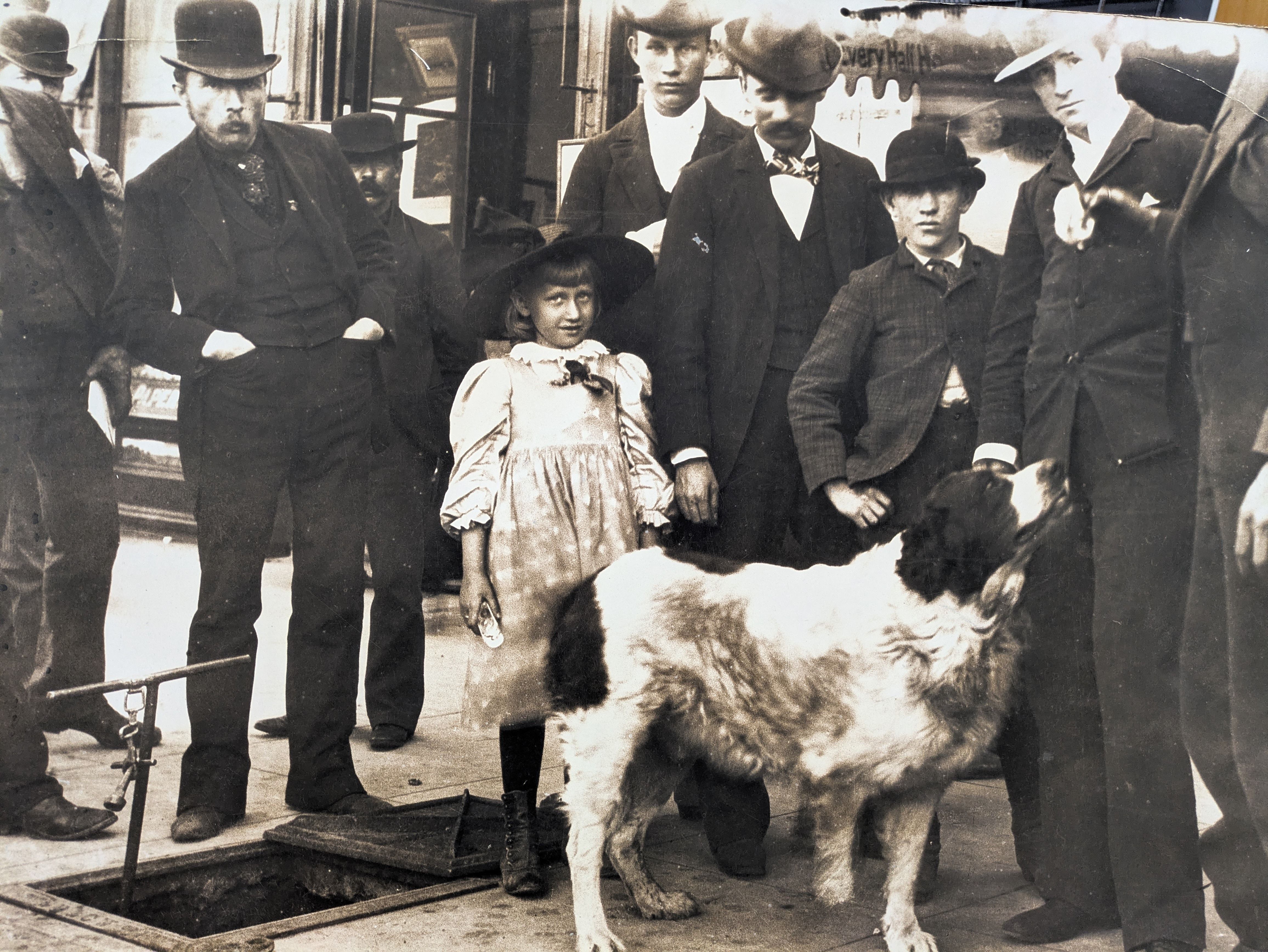 Black and white photo of a group of men wearing suits and hats, a young girl in a dress and hat, and a large dog standing on a city street near an open manhole.