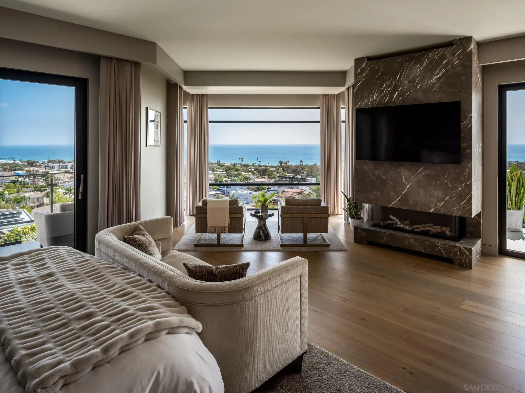 Luxurious bedroom with ocean view through large windows, beige sofas and curtains, a dark marble fireplace, and a wall-mounted TV; round coffee table and plants complete the space.