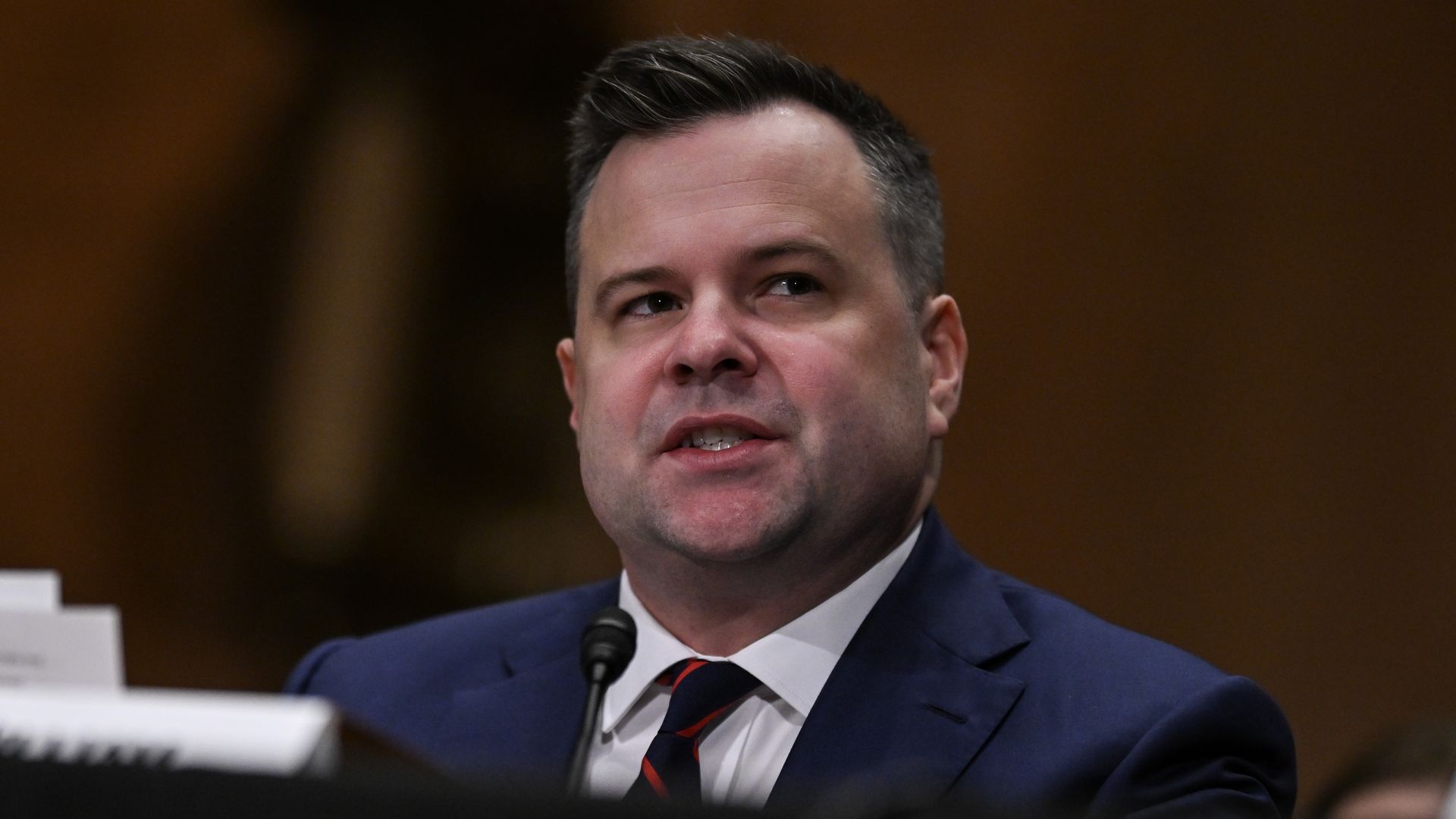 Bill Pulte wearing a blue suit and sitting in front of a wooden wall speaks into a microphone at a black table.