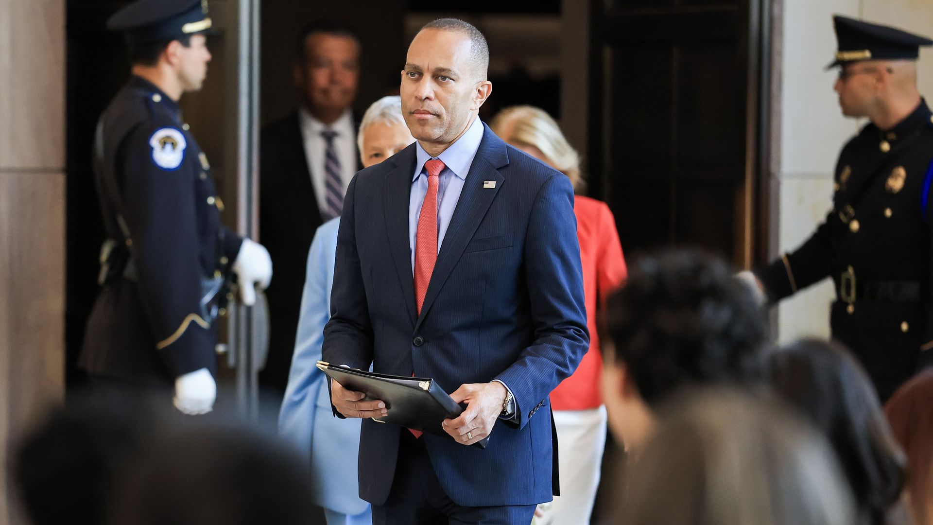 A man in a blue suit and red tie walks through a doorway, holding a folder. Two uniformed guards flank the entrance as blurred people look on.