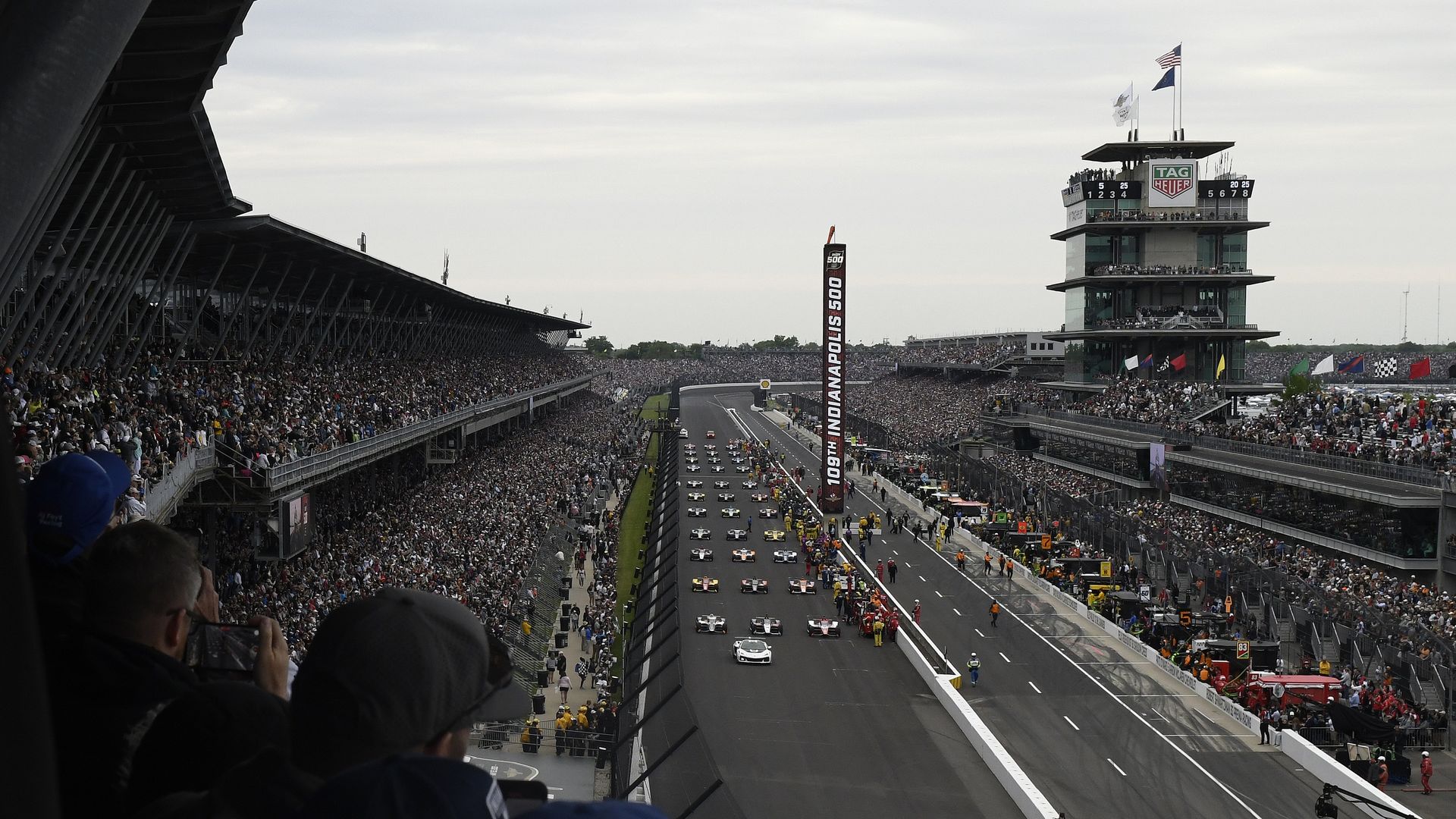 Wide view of a packed Indianapolis 500 race: left grandstands, right control tower with flags, and a busy pit/finish line with pace cars and banners reading "INDIANAPOLIS 500" along the center.