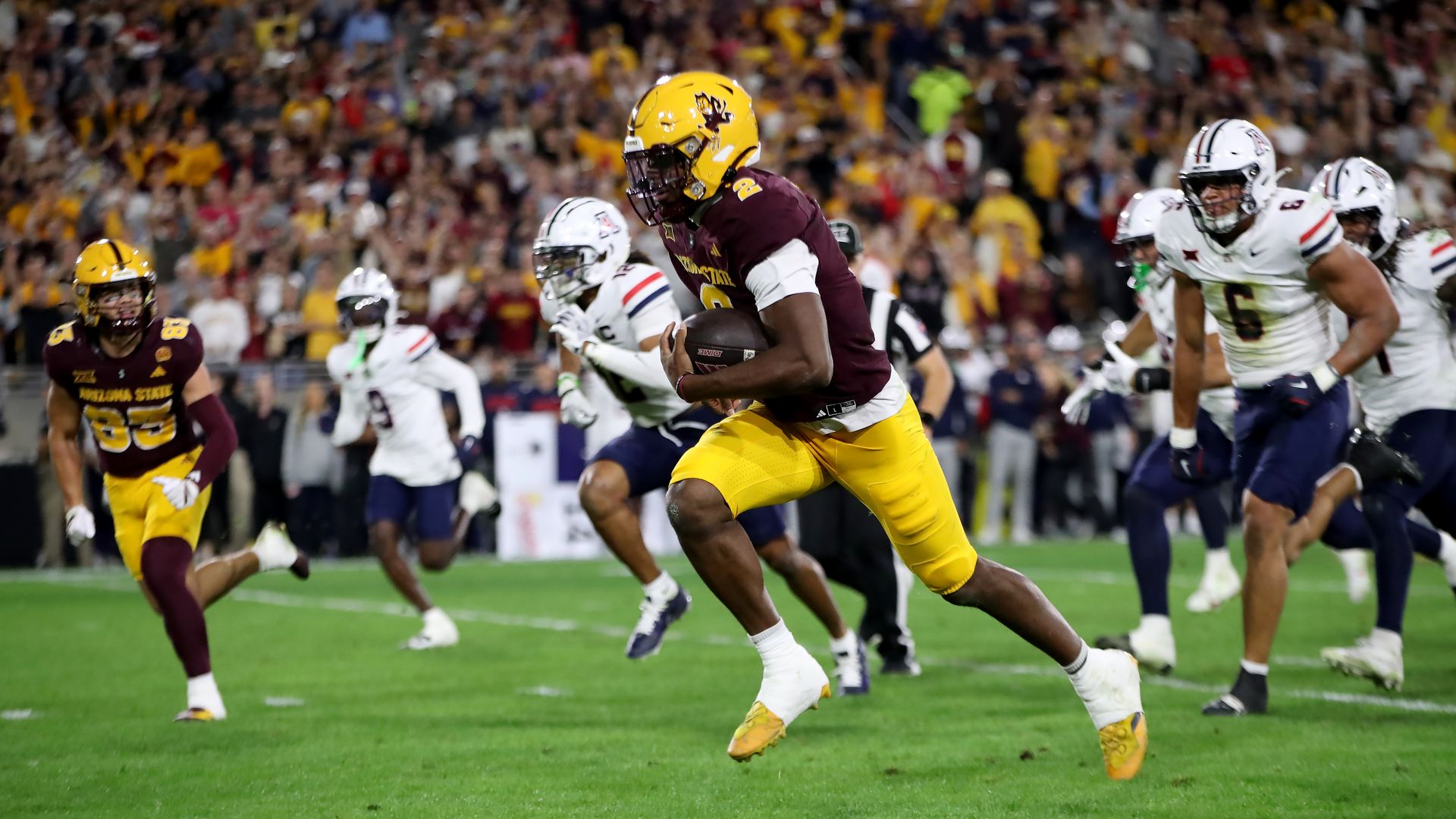 A football player in a yellow helmet and pants and a maroon jersey runs with the football while opposing players in white jerseys and helmets and blue pants chase him. 