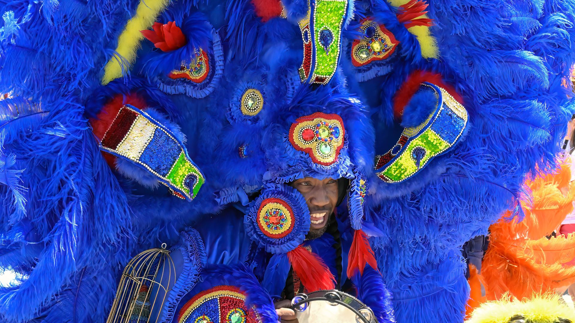 Big Chief Juan Pardo is seen smiling while performing music and wearing his Mardi Gras Indian suit. A beaded headdress of blue feathers surrounds his face.