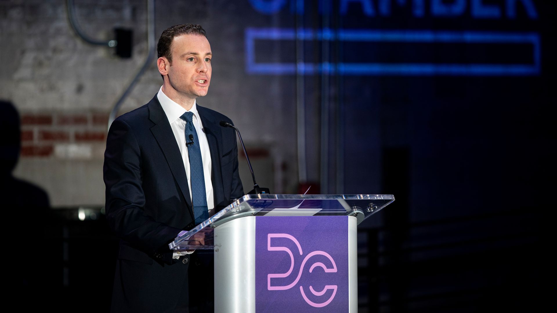 A man in a dark suit and blue tie gives a speech at a clear podium with a purple panel, against a dim industrial backdrop and a blue-lit sign that reads CHAMBER.