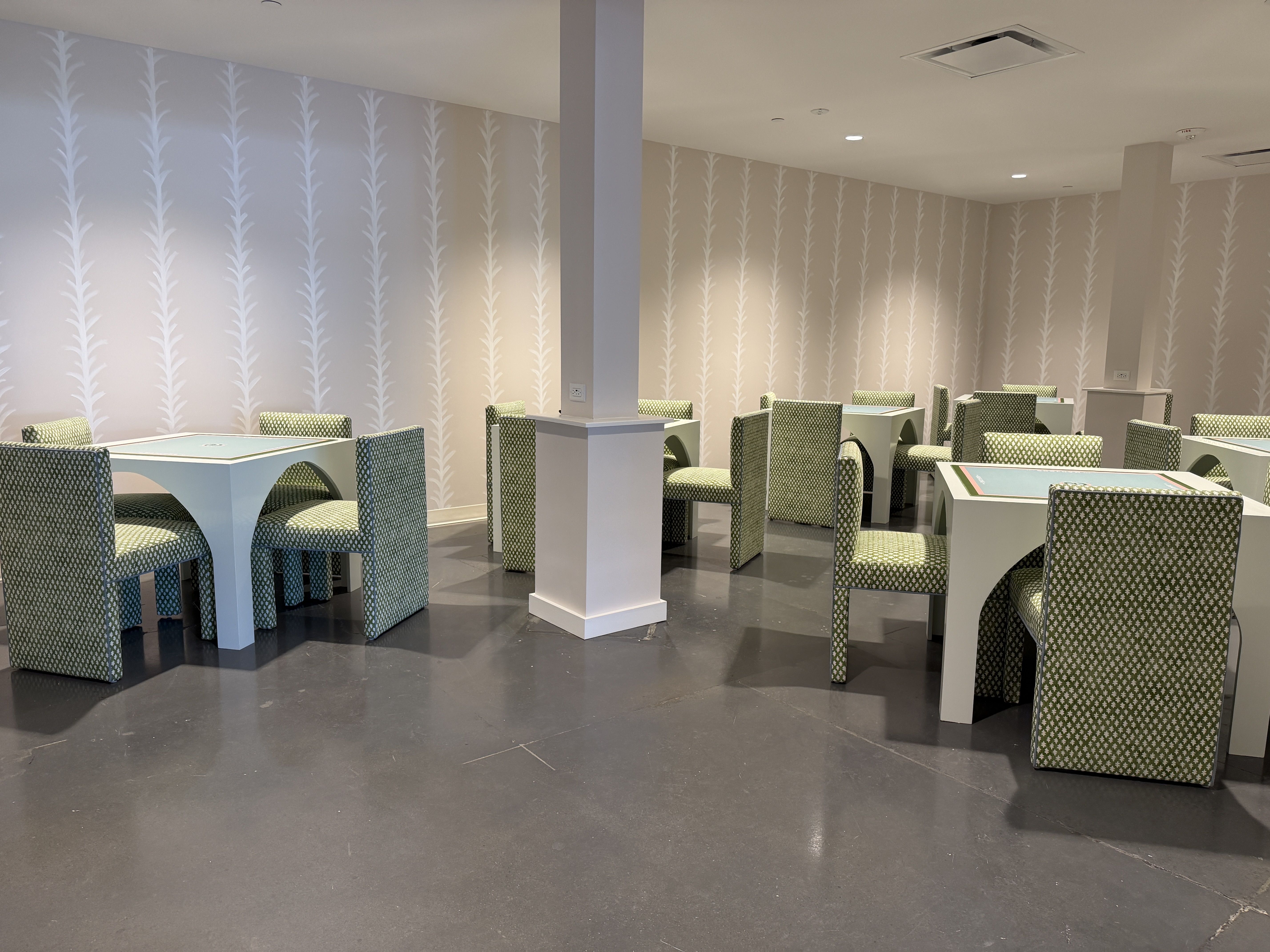 The Queen's Mahjery. An airy room with light grey walls covered in white vertical leaf-pattern wallpaper. Green patterned chairs surround white square tables; a central white pillar stands in the middle under soft overhead lighting.