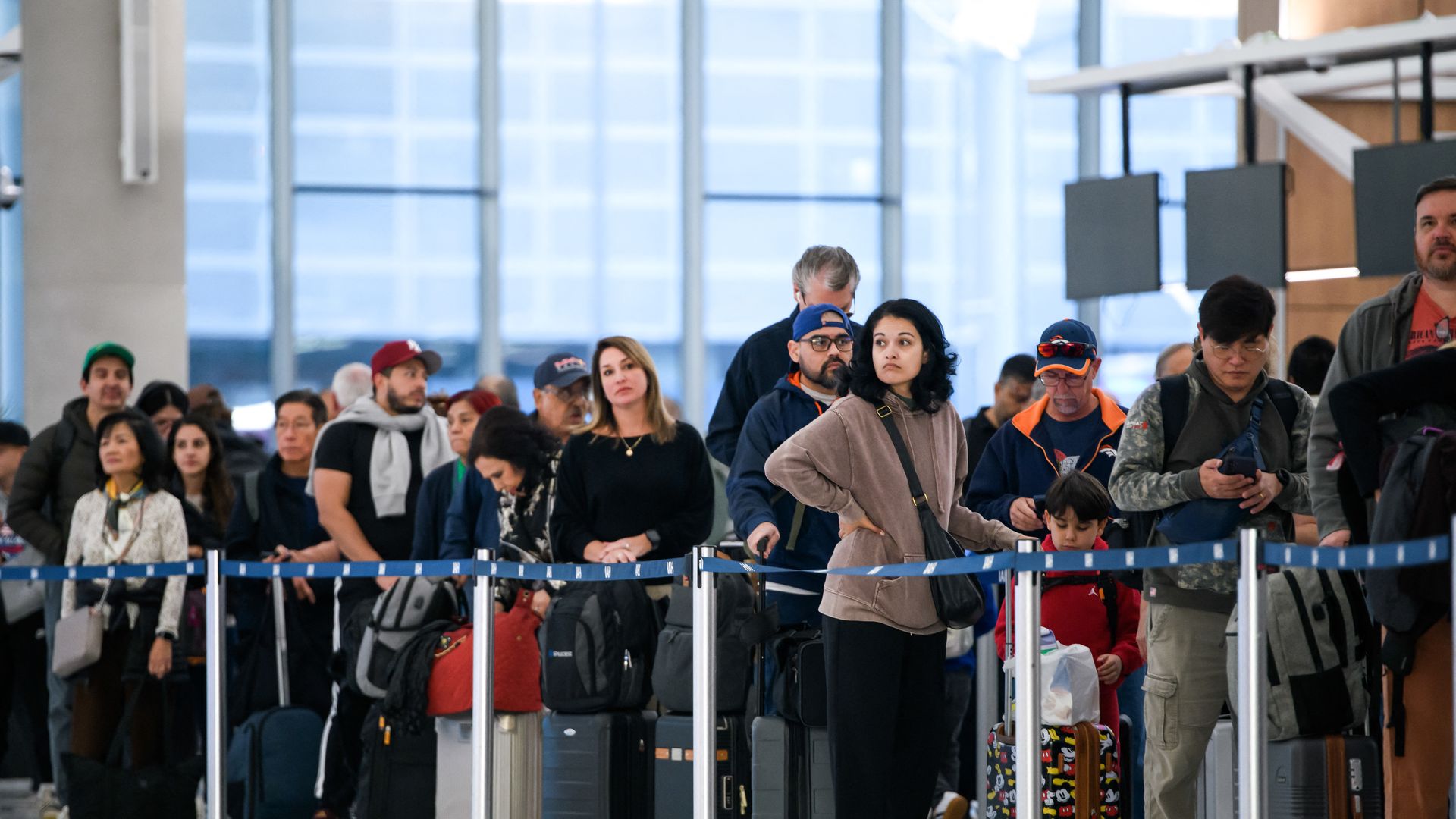 several people are standing in a long line at an airport, looking out while standing next to their luggage