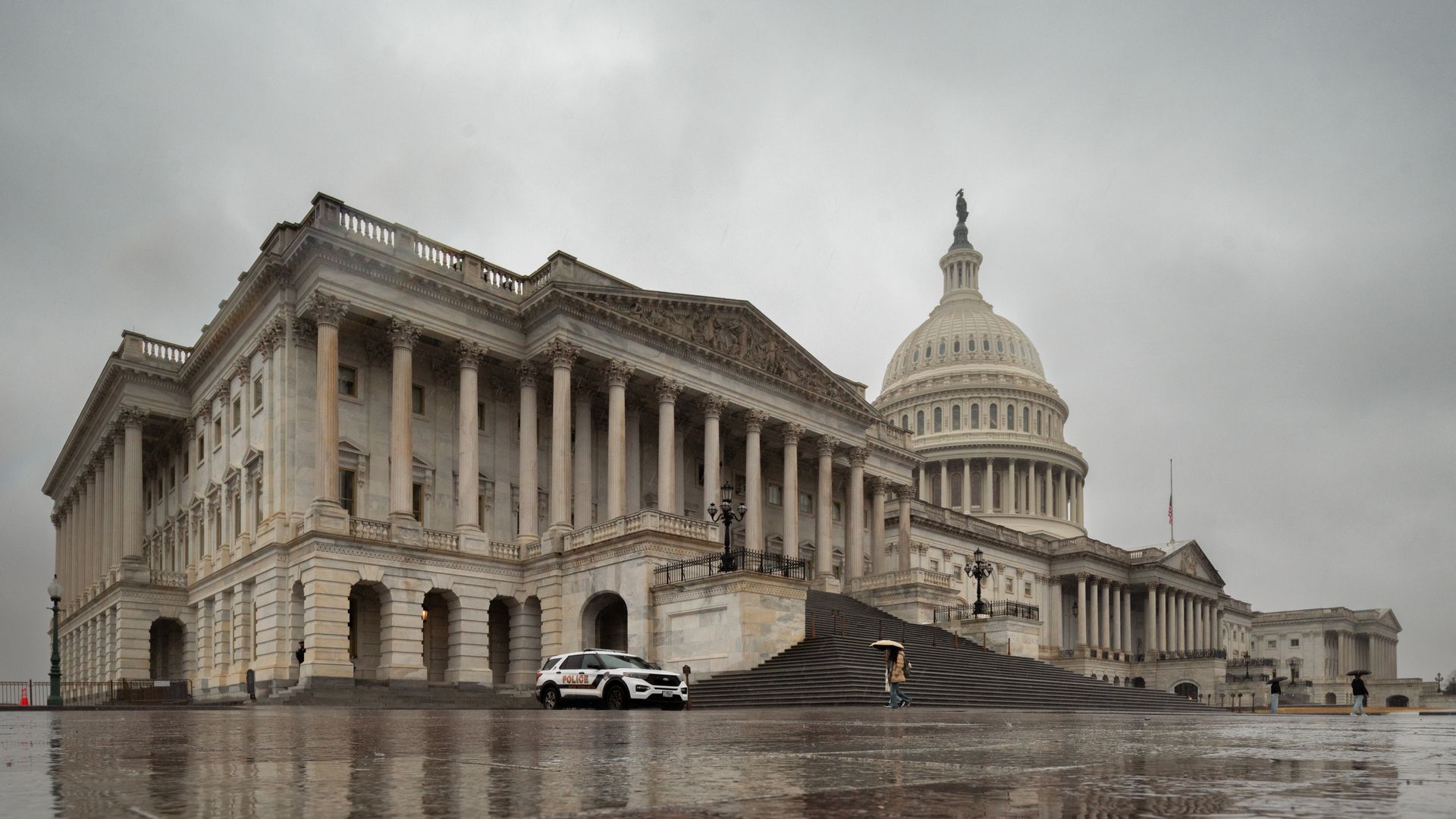 The House of Representatives side of the U.S. Capitol Building is seen on a stormy day in Washington, DC. A security SUV is parked out front and a person with an umbrella walks toward the steps. 