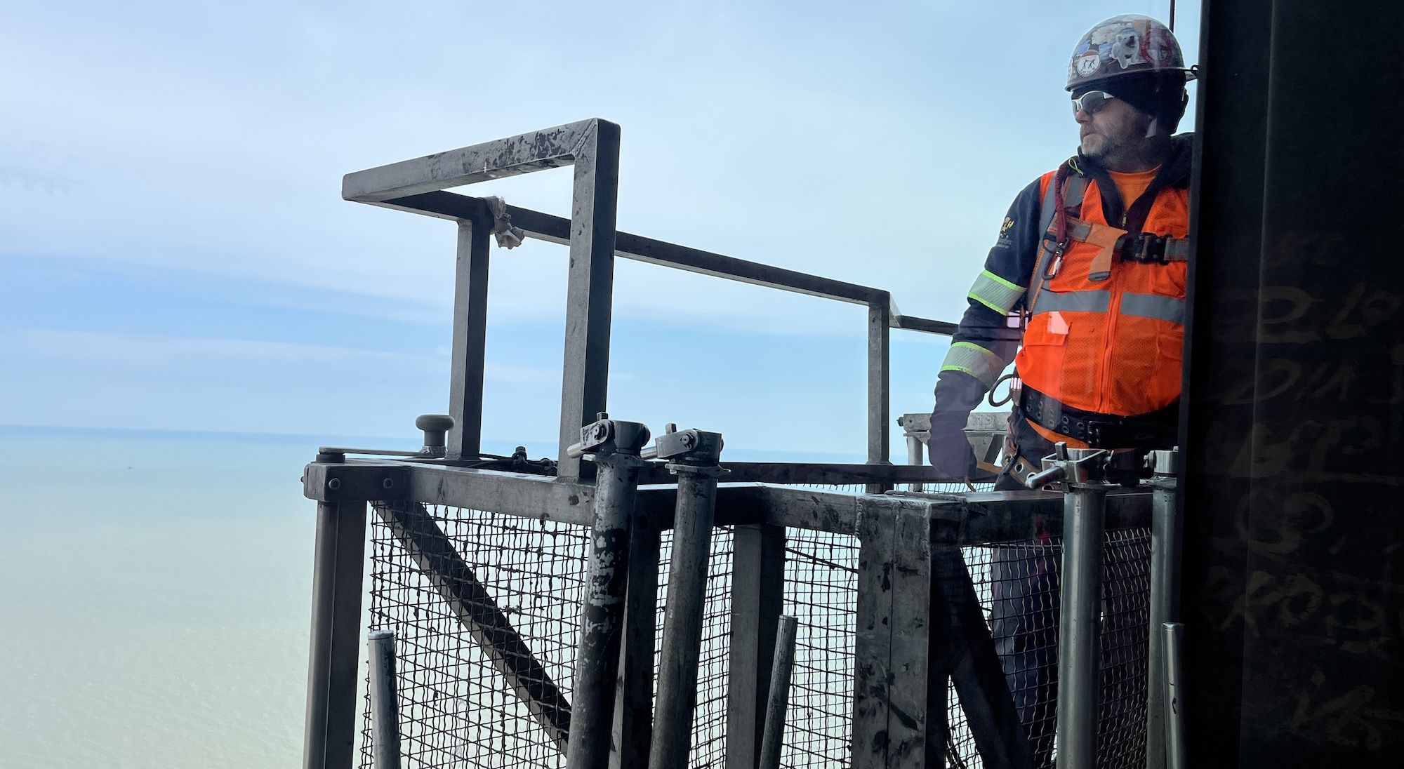 Construction worker in an orange safety vest, helmet with stickers, gloves and sunglasses stands on a weathered metal scaffold by the sea, with a clear blue sky in the background.