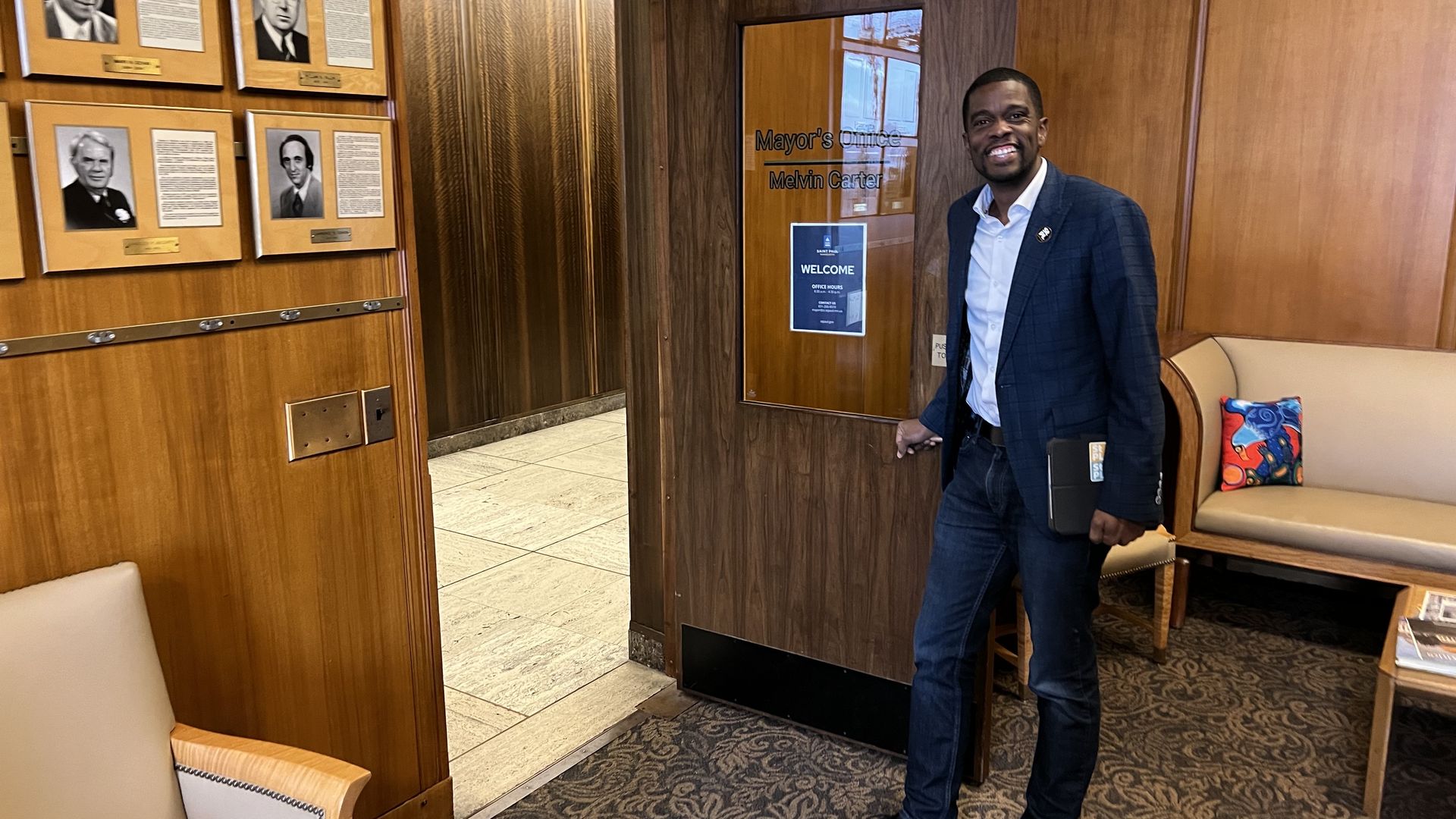 Man in blue blazer and jeans stands smiling at the open door of the Mayor's Office of Melvin Carter, with wood-paneled walls, portraits, beige chairs, and colorful pillow visible.