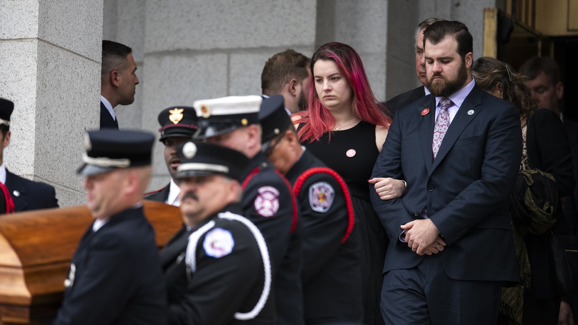 Funeral scene outside a gray stone building: uniformed pallbearers carry a wooden coffin; a woman with pink hair and a suited man stand solemnly as others look on.