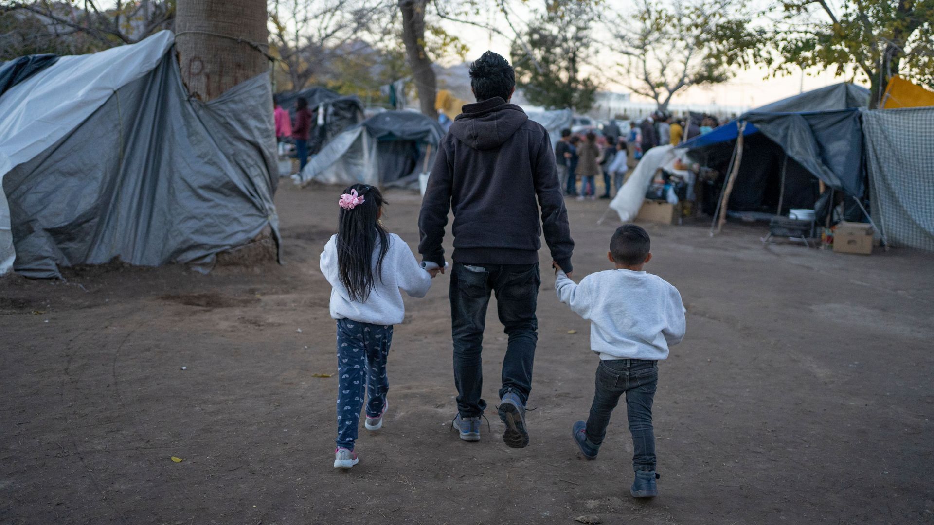 Photo of a man walking while holding the hands of two children, one on each side, at a temporary camp