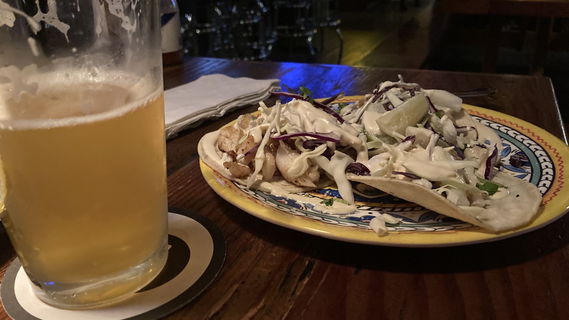 A half-full pint of beer next to a yellow-rimmed plate holding three soft tacos.