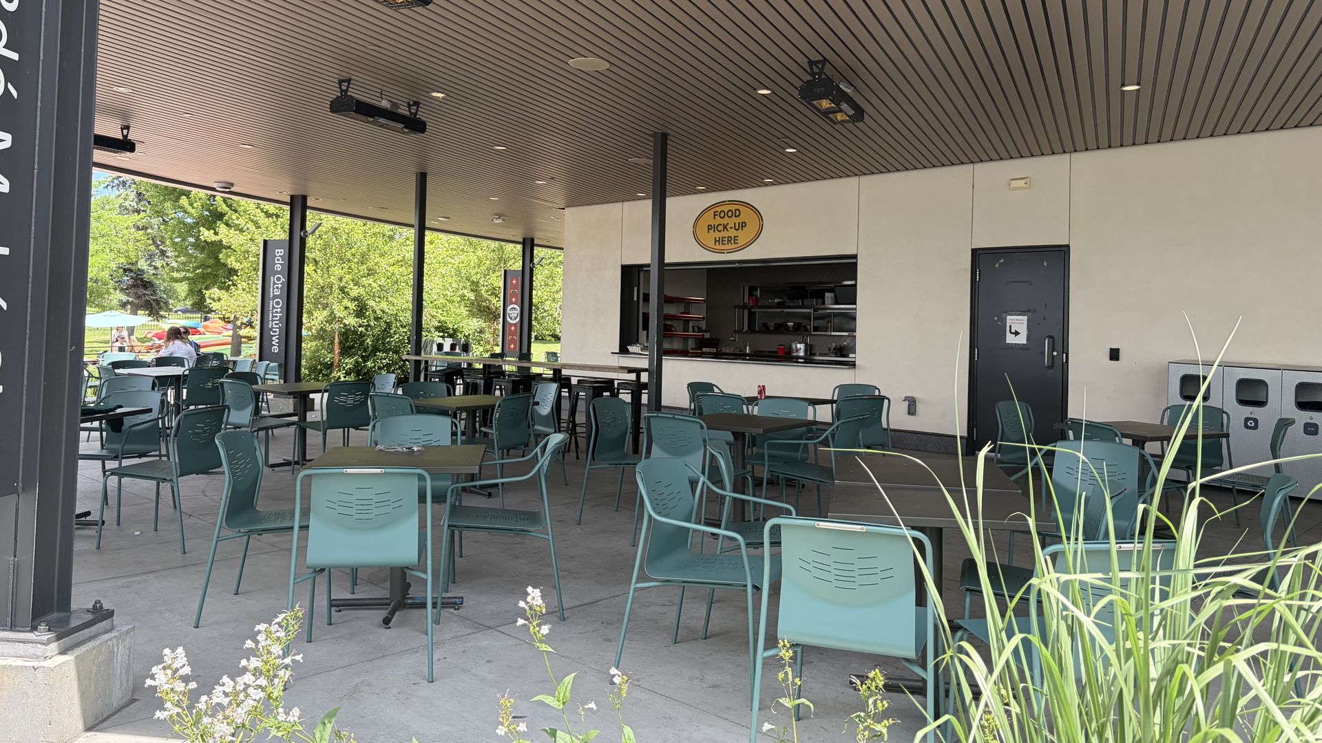 Outdoor cafe seating under a brown slatted ceiling with teal chairs and many tables. A service counter on the right and a yellow "Food Pick-Up Here" sign; trees and people in the background.