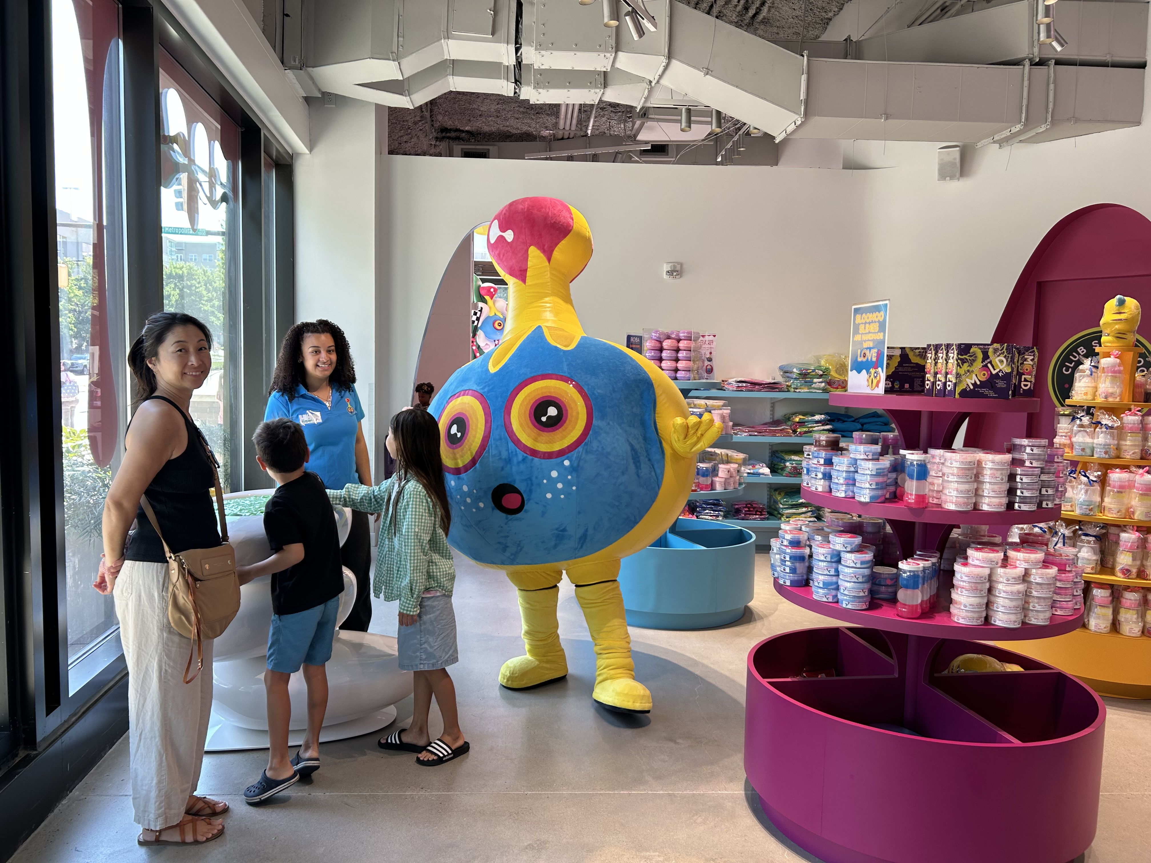 Inside a bright toy store, a large blue mascot with a pink and yellow head waves at adults and kids while colorful shelves of candy and merchandise surround them; windows are on the left.