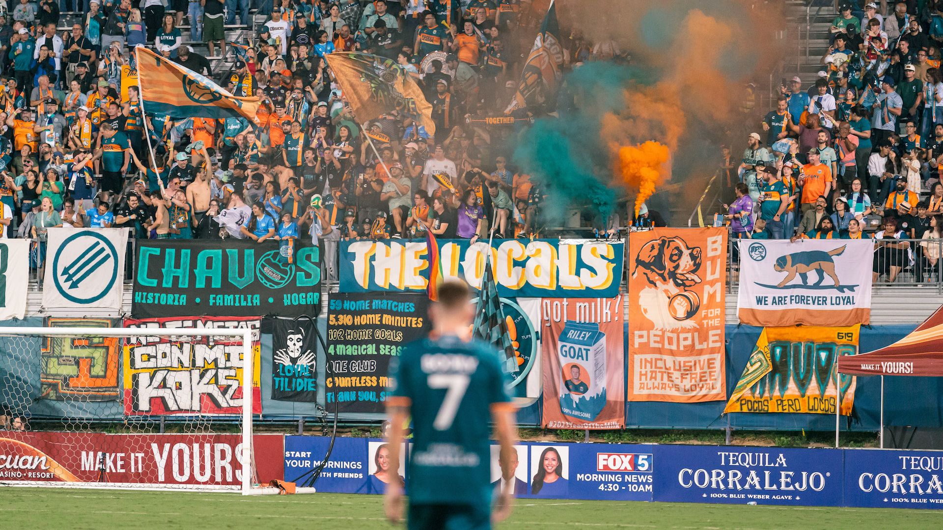 A soccer player out of focus stands on the field facing a crowd of fans in a stadium.