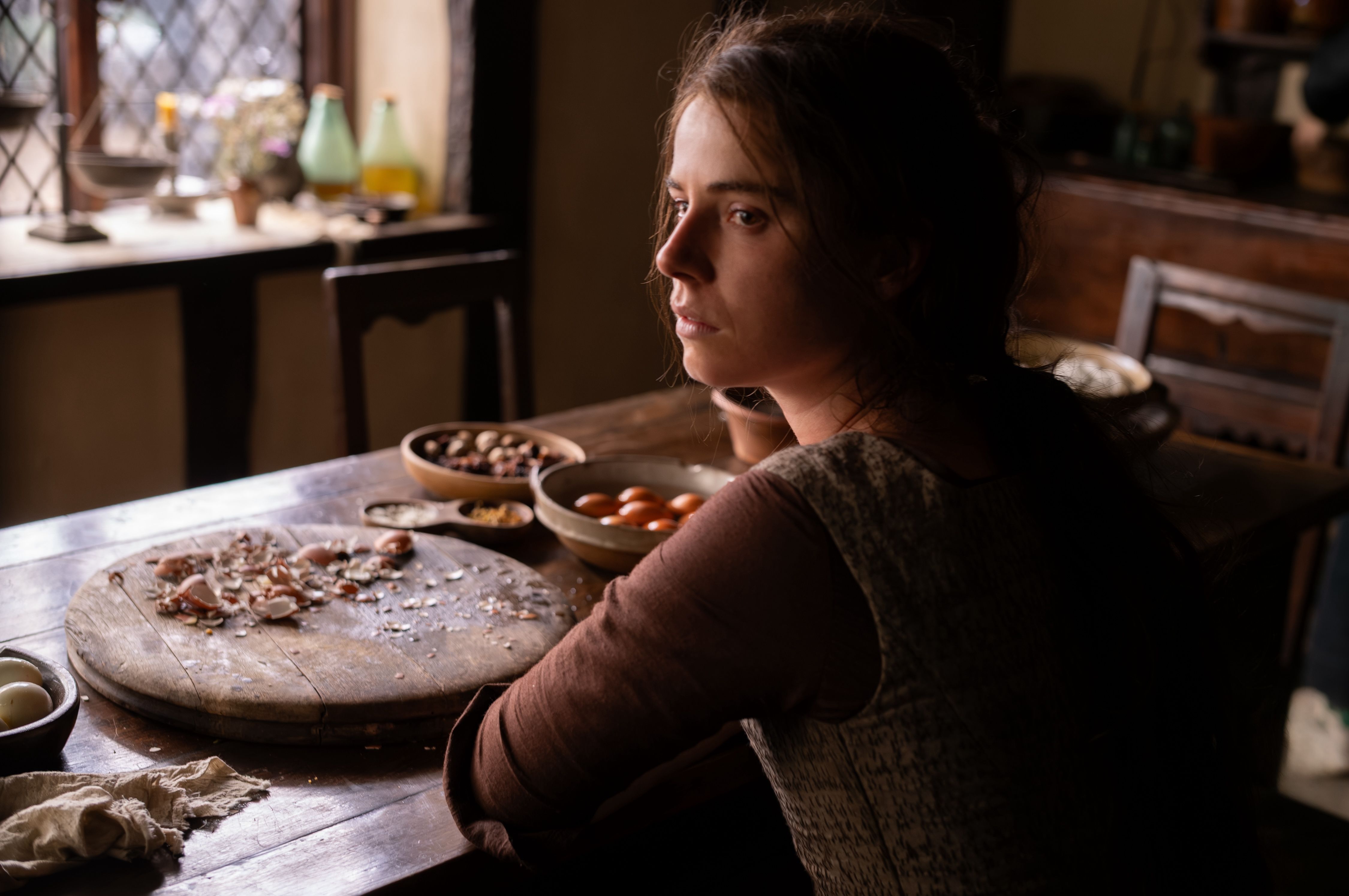 A woman sits at a wooden kitchen table, looking off to the right. Bowls of nuts and eggs, a round cutting board with eggshells, and scattered shells fill the scene under warm light.
