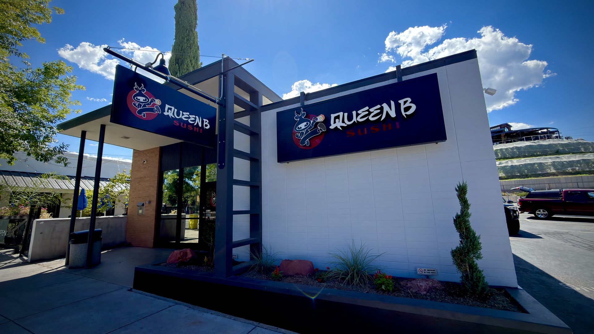 Bright sunny day at a modern sushi restaurant with dark blue signs reading "QUEEN B SUSHI" and a cartoon bee logo; tall cypress tree behind, blue sky with white clouds, and a parking lot to the right.