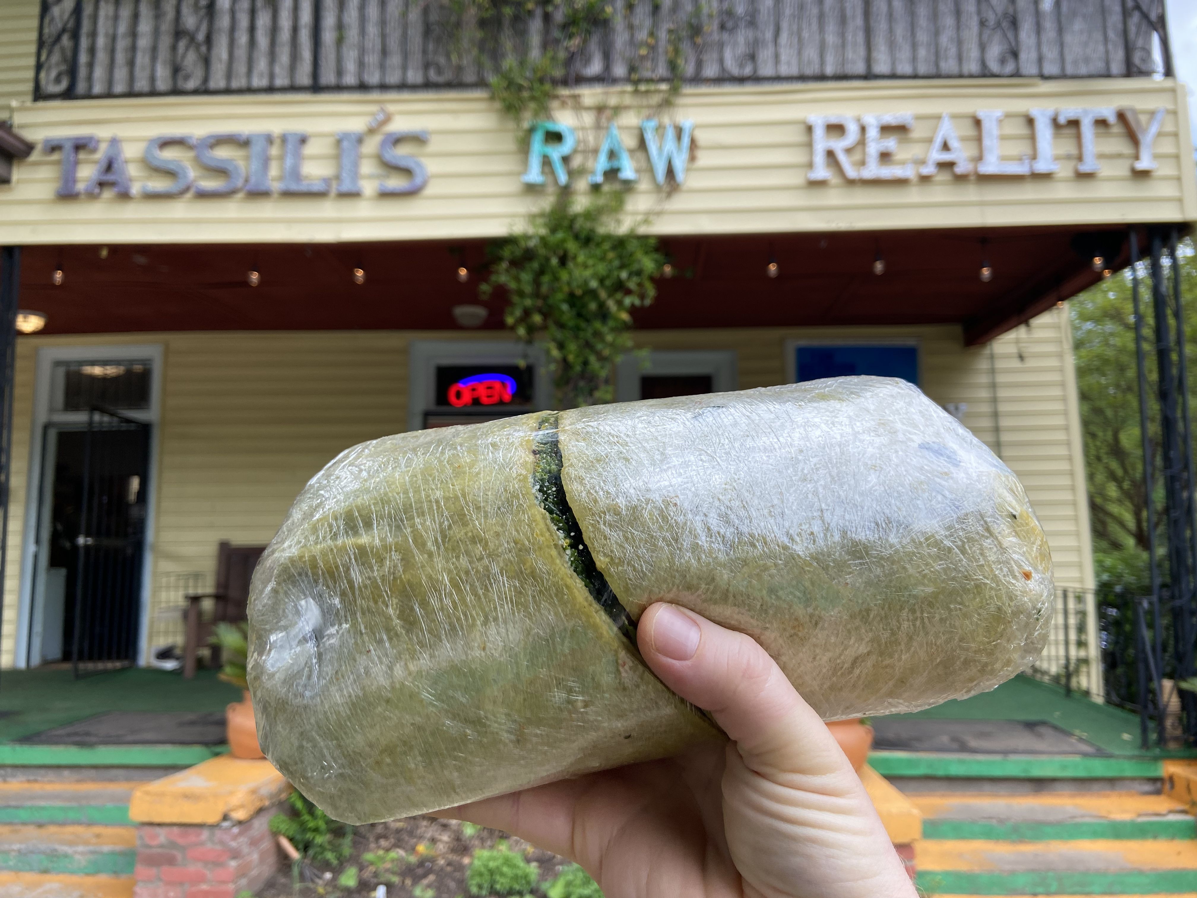 A hand holds a massive spinach wrap filled with raw food in front of Tassili’s Raw Reality