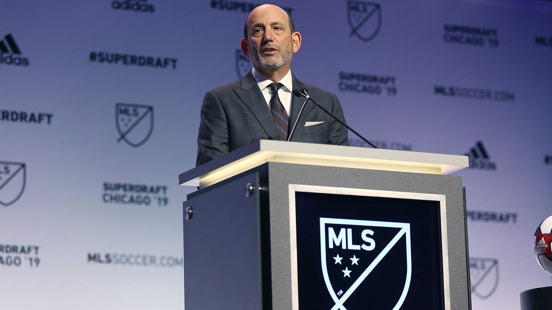 In this image, Don Garber stands in a suit and speaks from behind a podium. 