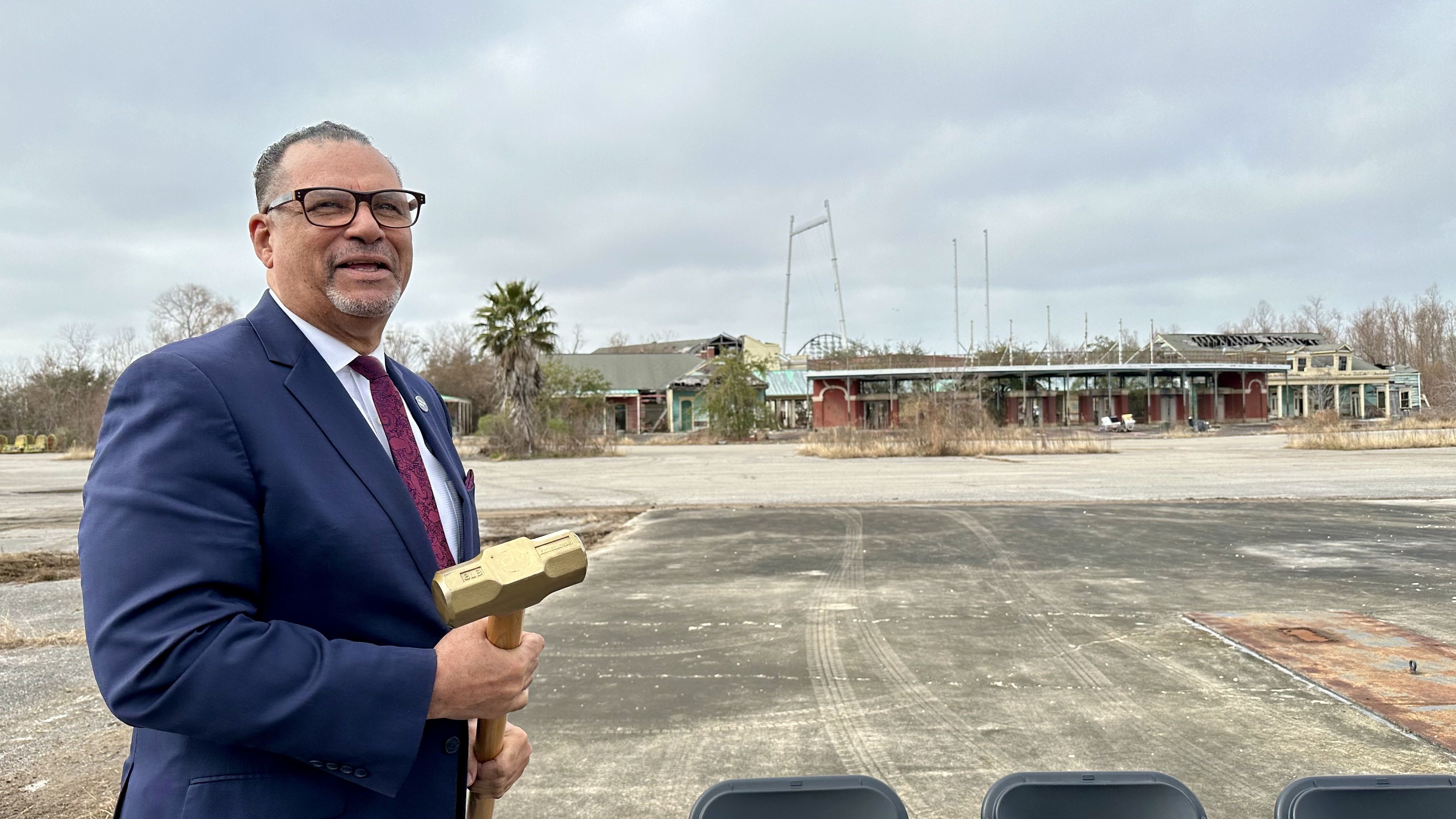 Man in blue suit and glasses holding a sledgehammer, standing outdoors on a cloudy day in front of an abandoned, rundown amusement park.
