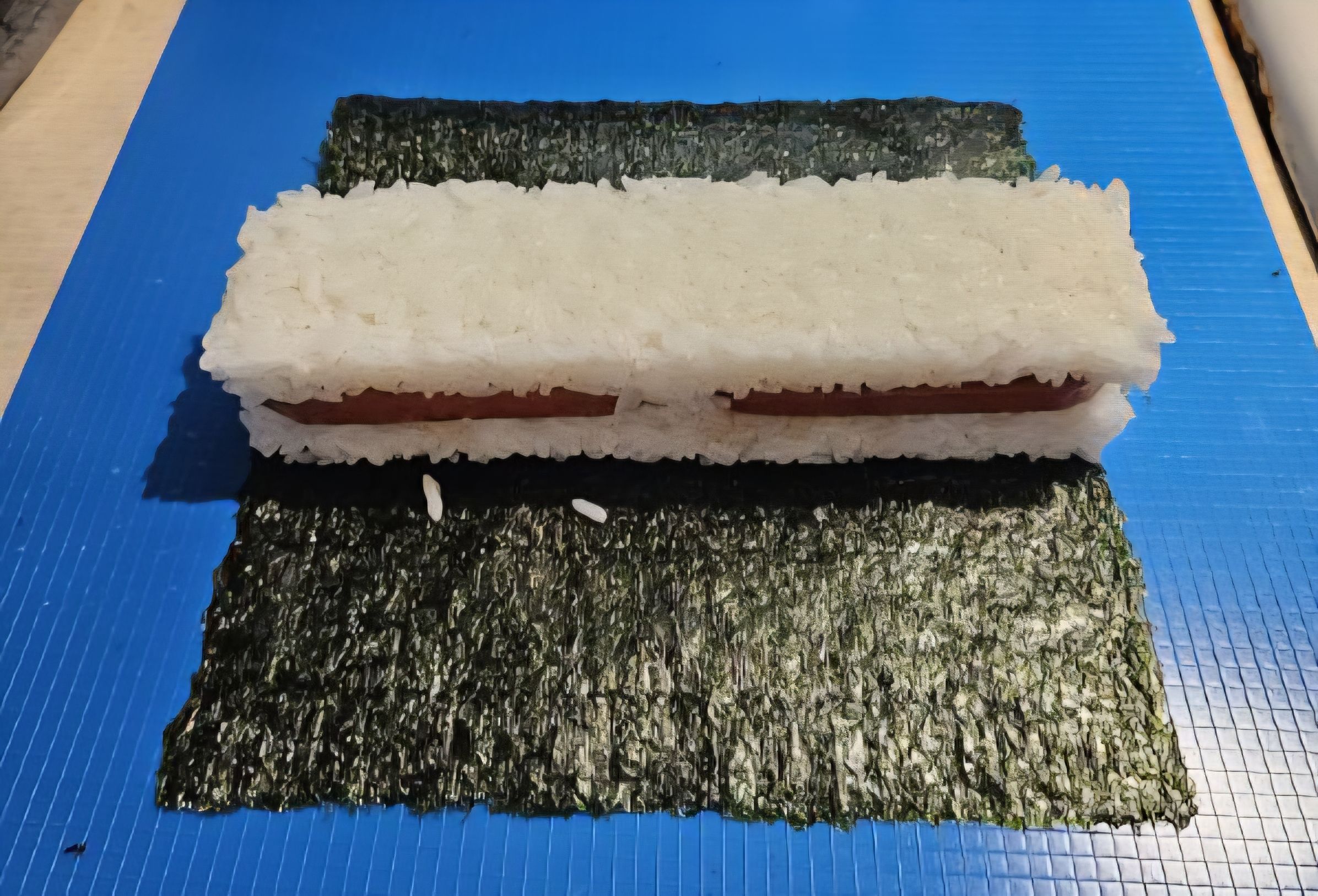 Rectangular sushi roll with white rice and a strip of meat, placed on a square sheet of dark green seaweed on a blue surface.