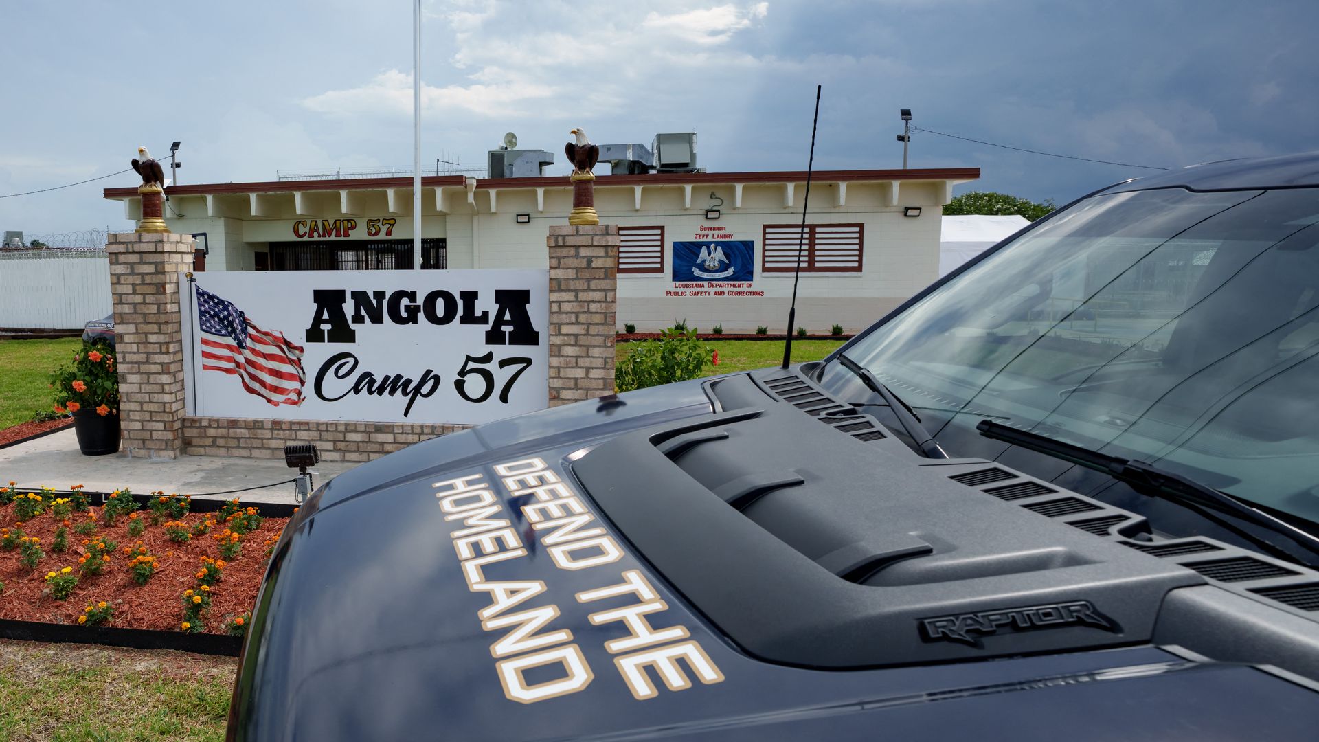 A car is parked directly in front of a sign for "Angola Camp 57" and a building with the same signage behind it. The hood of the car says "Defend the homeland."