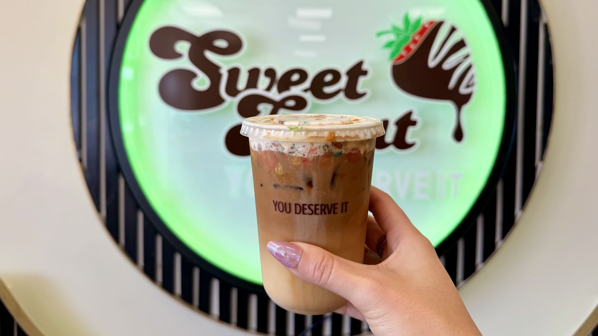 Hand with purple nails holds a tall iced coffee cup with a clear lid and colorful ice, in front of a glowing green circular sign that reads "Sweet"; the cup shows the phrase "YOU DESERVE IT".