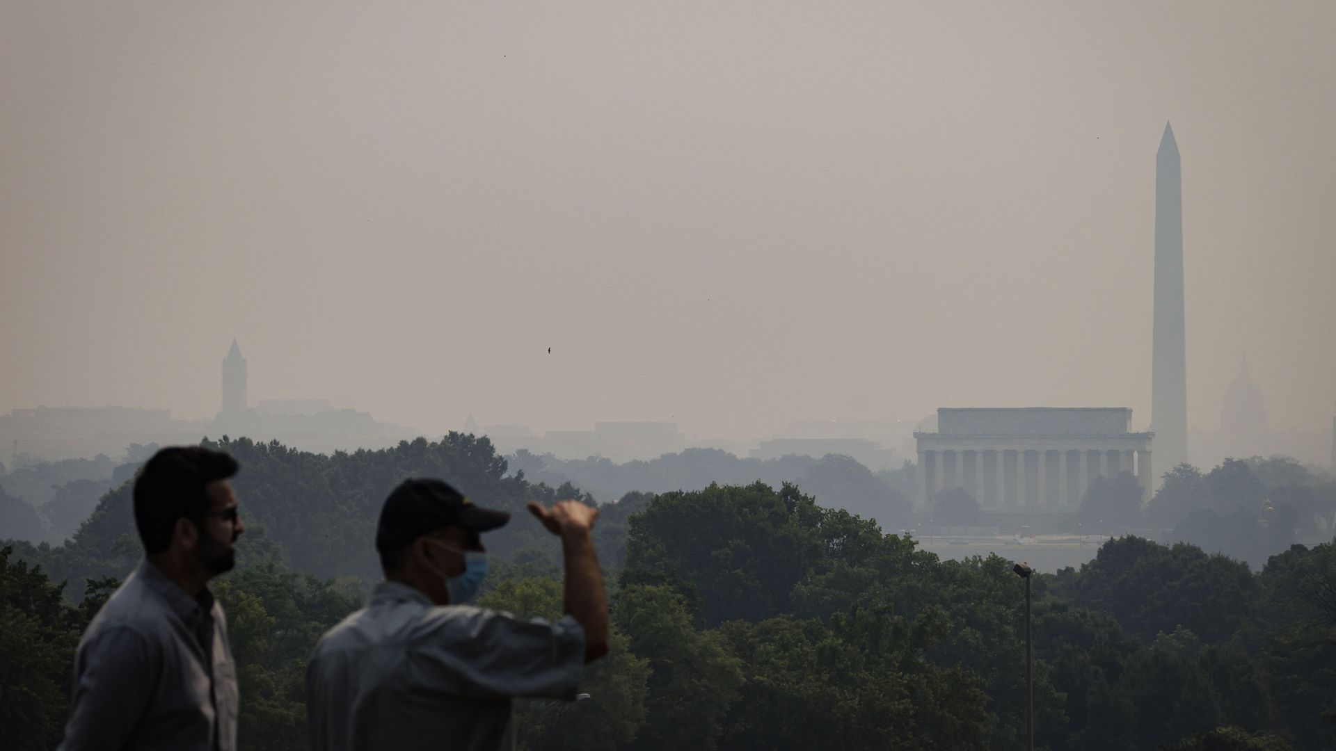 Two people, one wearing a mask outdoors, are photographed with hazy skies in the background and partially obscured Washington monuments