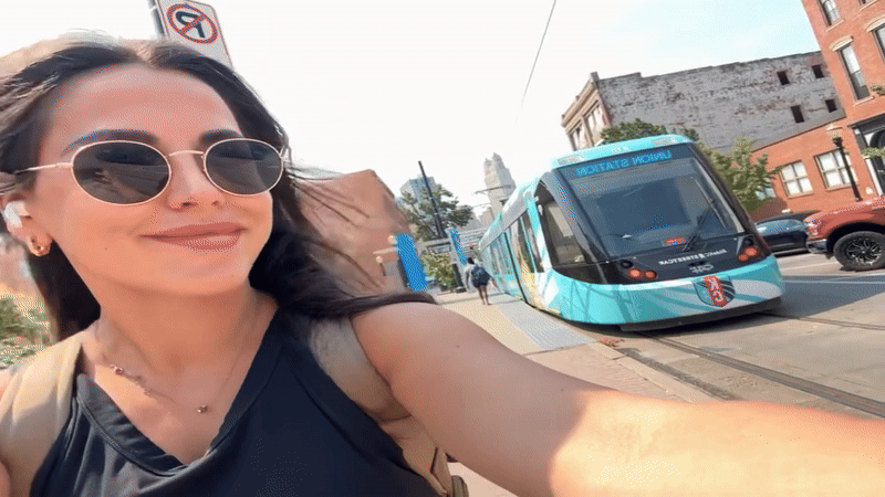 A woman wearing sunglasses and a black top takes a selfie outdoors near a modern blue streetcar on a sunny day in an urban area with buildings and a no parking sign visible.