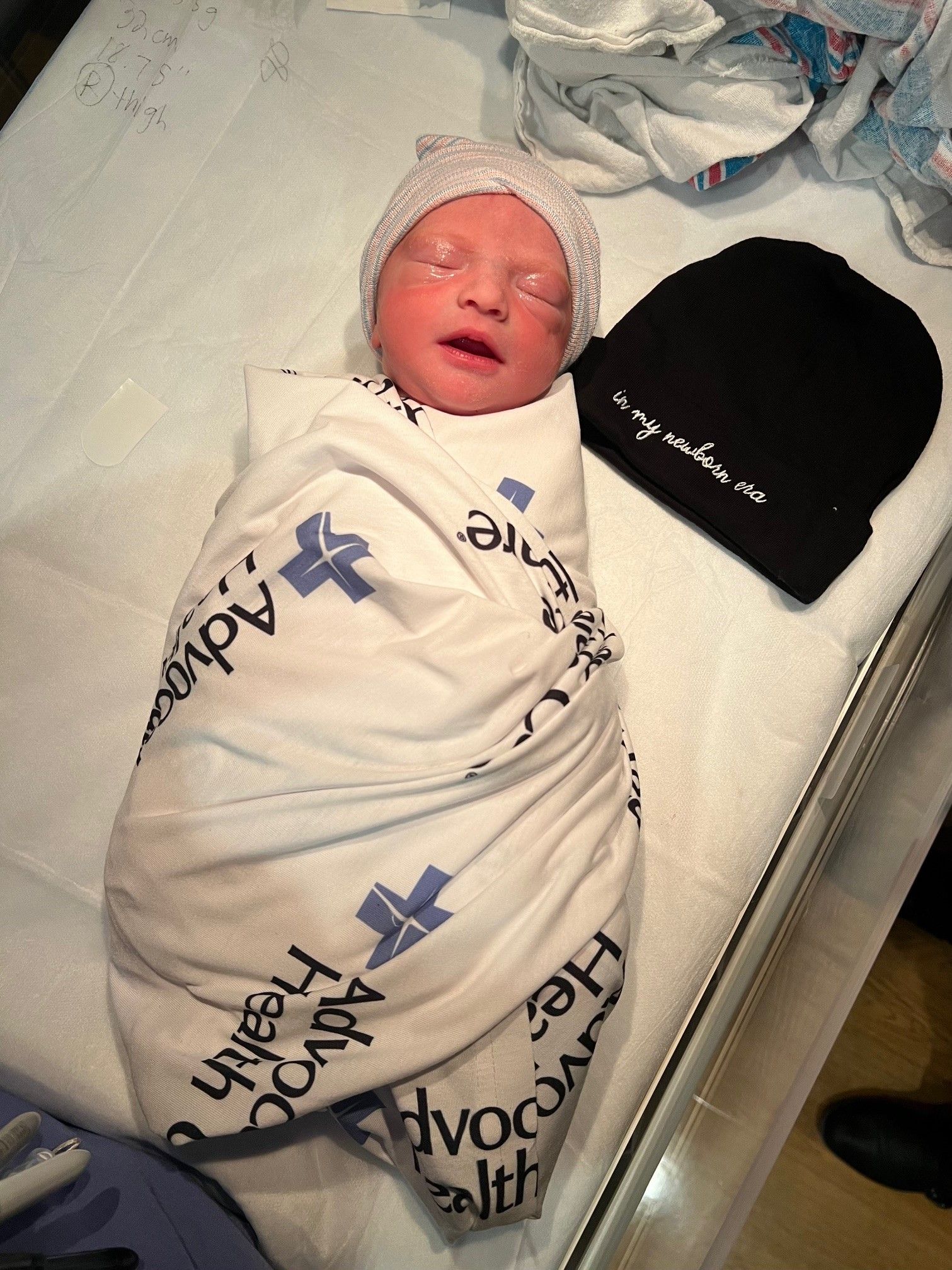A photo showing a newborn baby wrapped in a swaddle with their eyes closed next to a beanie that says "in my newborn era."