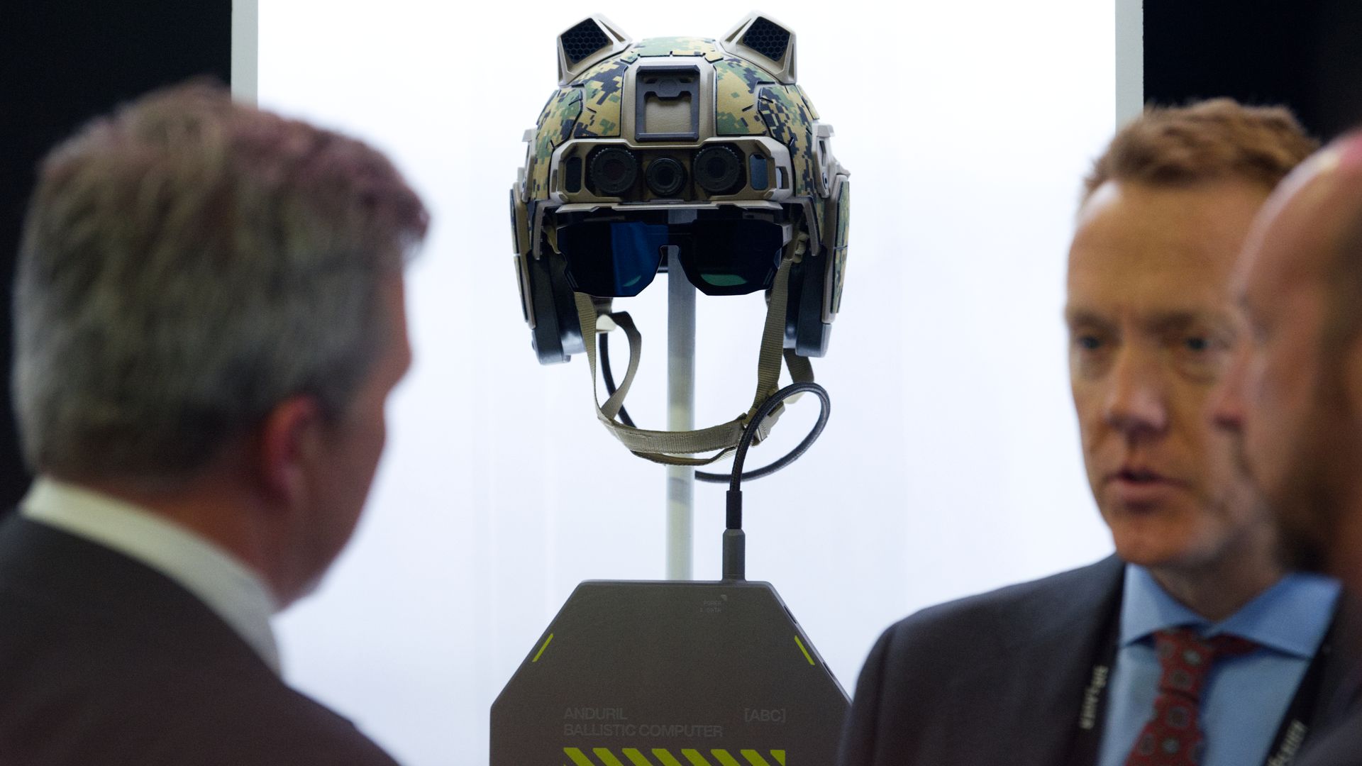 High-tech military helmet with camouflage pattern and attached sensors, displayed on a stand labeled "Anduril Ballistic Computer," with three men in suits talking nearby.
