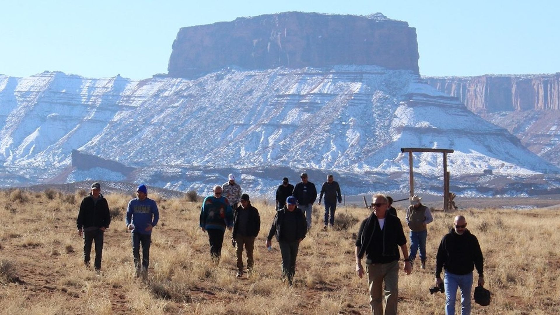 A film crew walks in front of a redrock mesa covered in snow.
