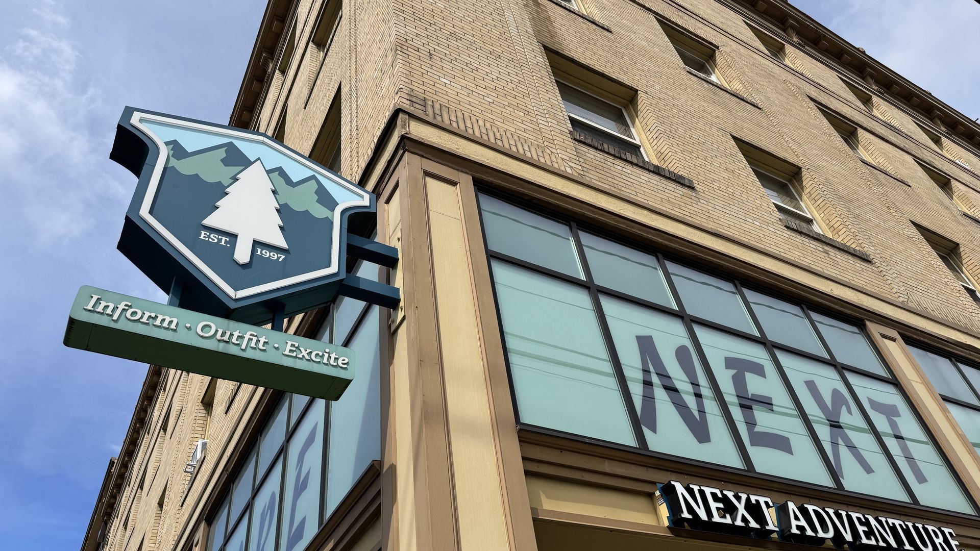 Corner of a beige brick building with a blue and green sign showing a white tree and text "EST. 1997" above the words "Inform · Outfit · Excite" and large windows with "NEXT" letters, and a "NEXT ADVENTURE" sign below.