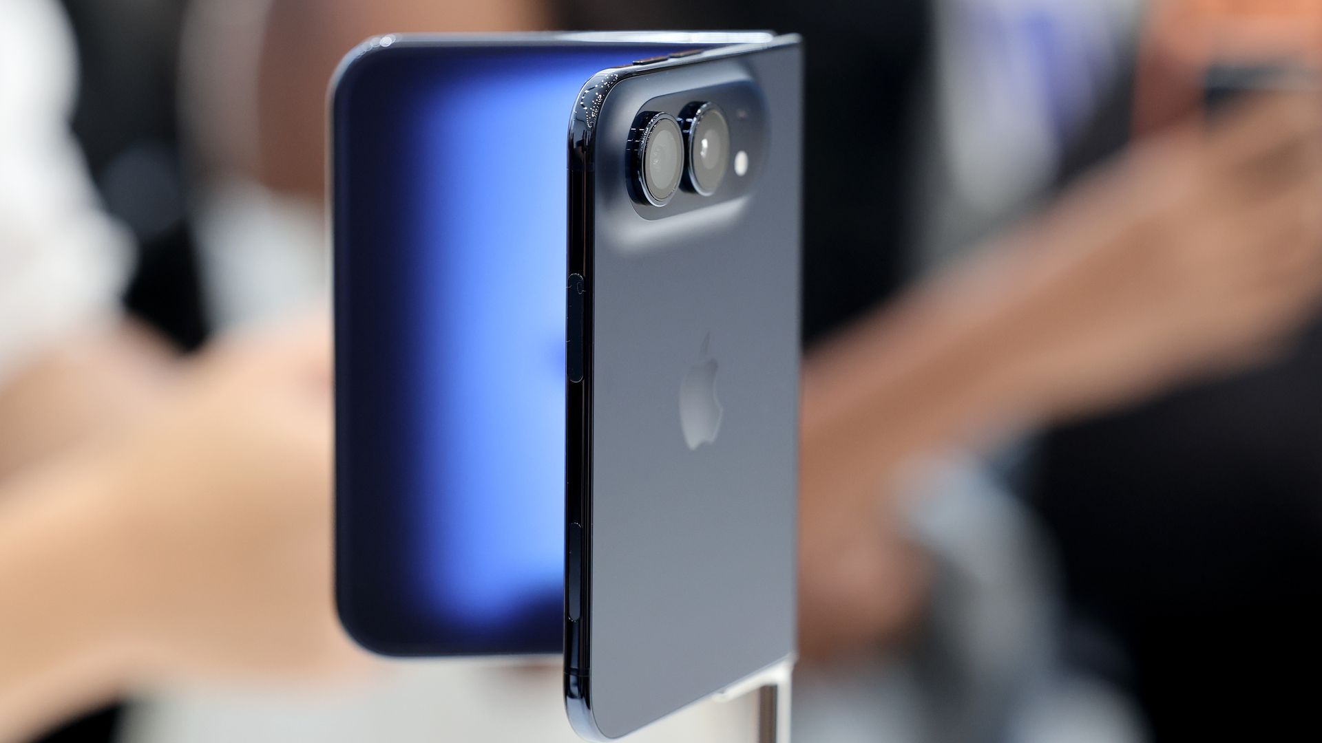 Close-up of a blue iPhone back showing a dual-camera bump with two lenses, glossy edges, mounted on a display with a blurred background.