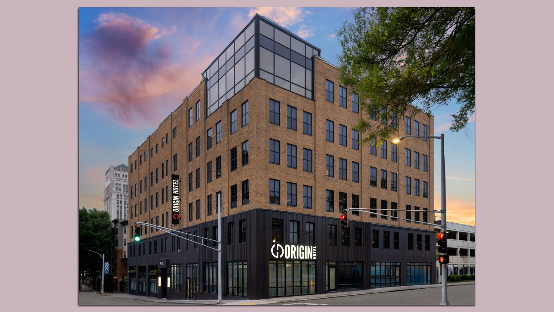 Exterior of a modern Origin Hotel at dusk with brick upper floors, black lower facade, large windows, streetlights, traffic signals, and a tree branch overhead against a colorful sky.