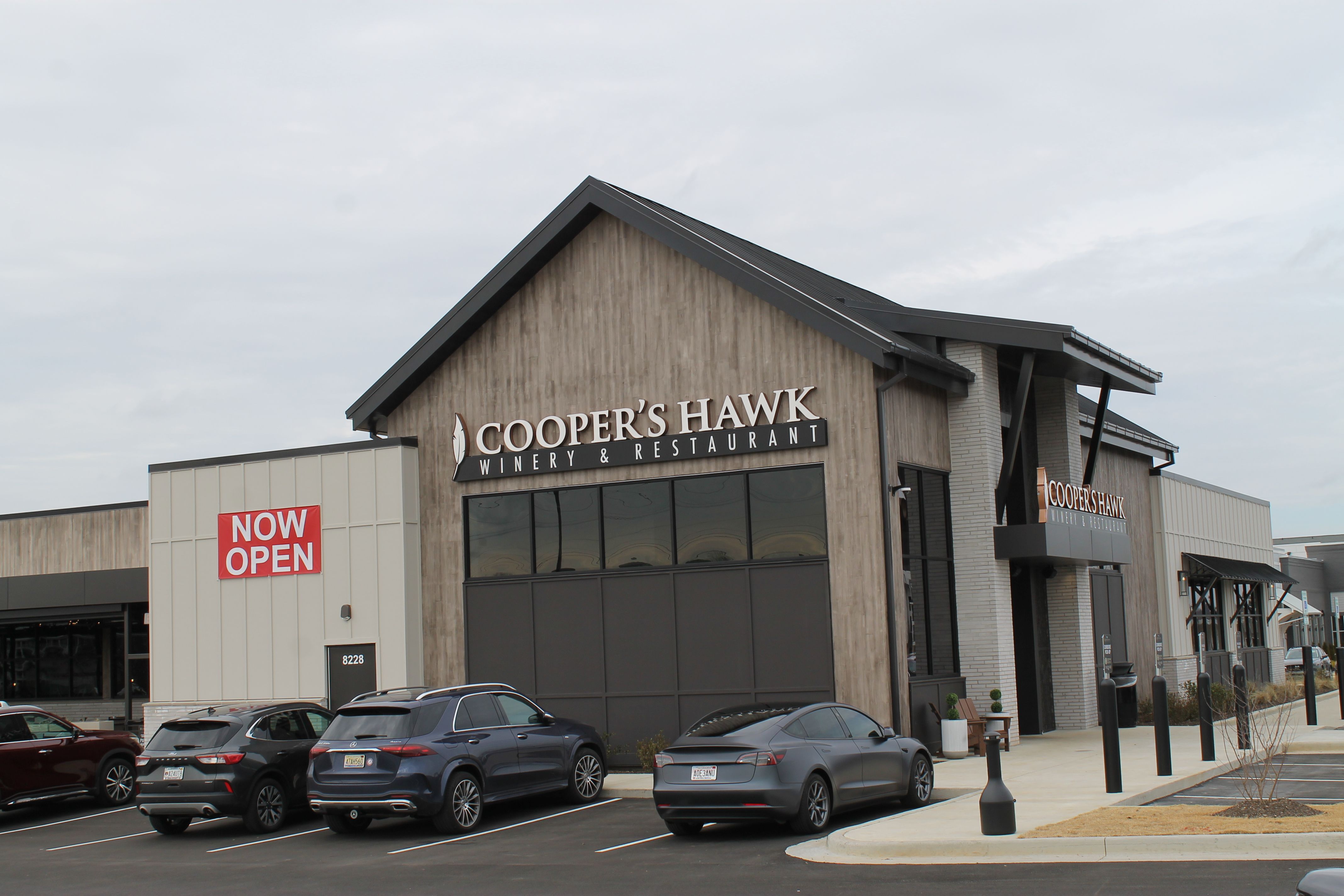 Exterior of Cooper's Hawk Winery & Restaurant with gray and wood facade, three parked cars, and a red "NOW OPEN" sign against an overcast sky.
