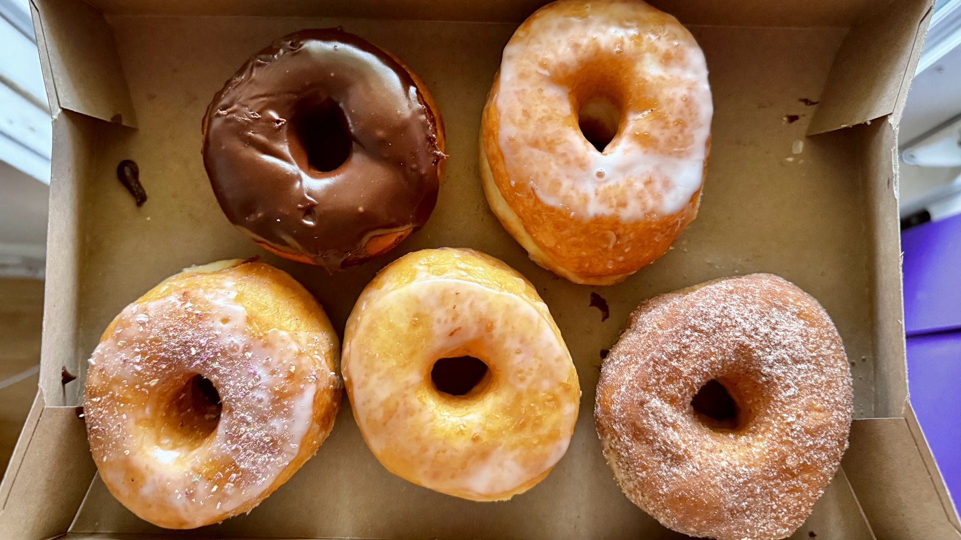Six donuts in a brown box: top row left to right—chocolate-glazed, white-glazed, and plain glazed; bottom row left to right—pink-sugar, glazed, and cinnamon-sugar.