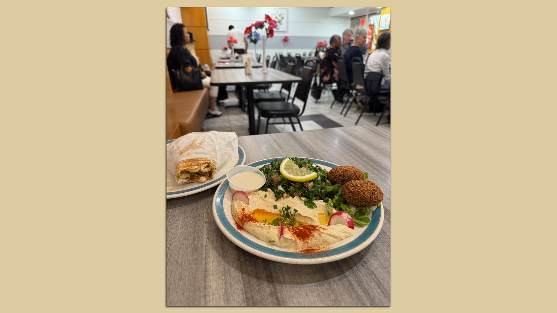 Close-up of a meal on a gray table: plate of hummus with paprika, lemon wedge, greens and falafel, plus a wrapped sandwich on the side and a small sauce cup. In the background, diners sit at tables in a casual restaurant.