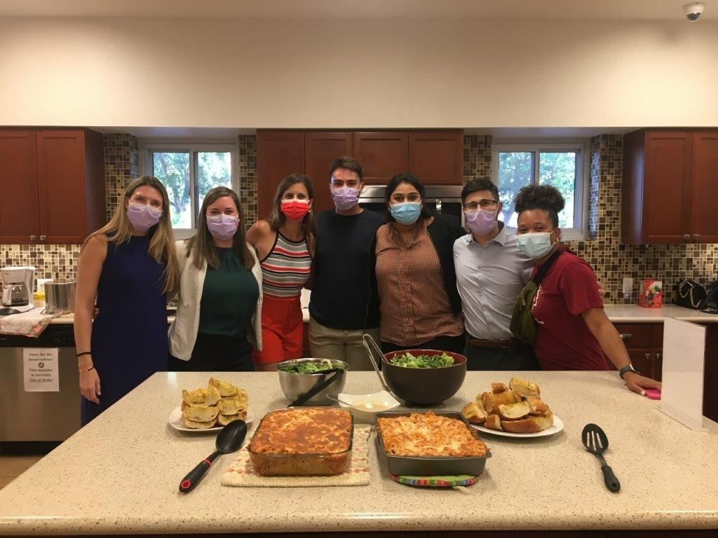 A group of people in masks stand around lasagna and salad in a kitchen.