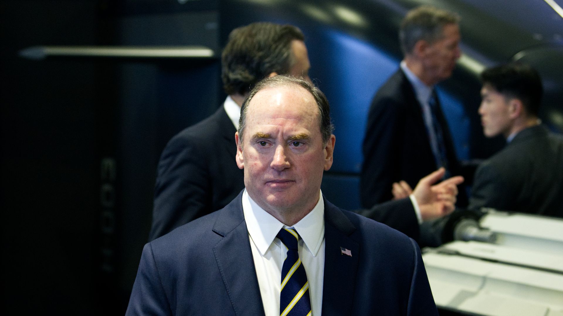Man in a navy suit, white shirt, and striped tie stands in front of a dark background with blurred people and objects behind him.