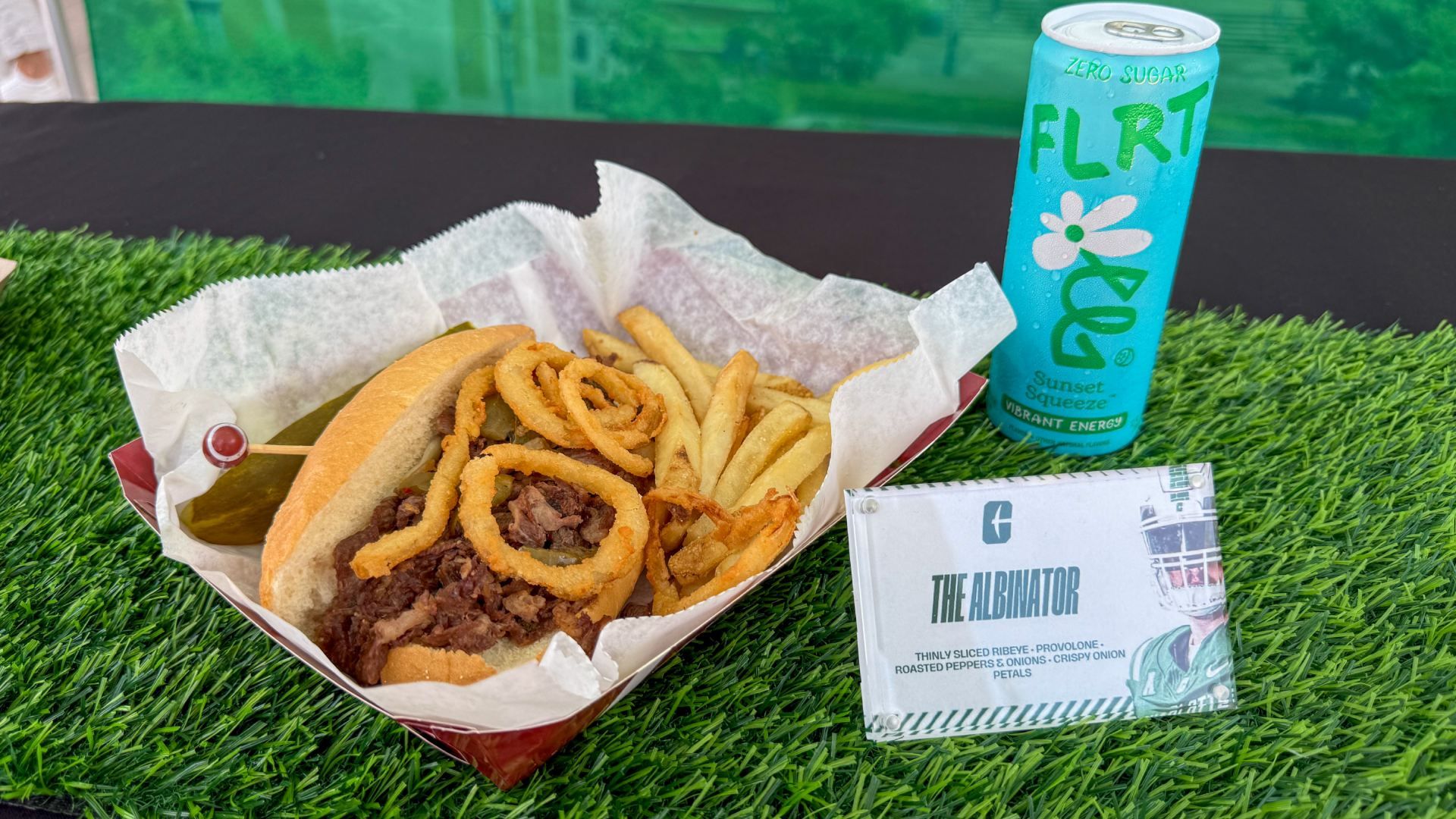 Fast-food basket on green artificial grass: beef sandwich with onion rings and fries, pickle spear, blue can labeled "FLRT" Zero Sugar, and a white "The Albinator" box.